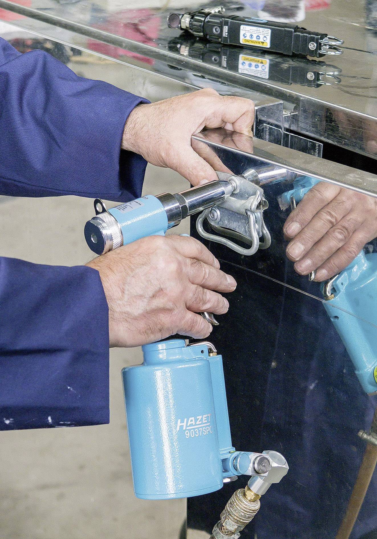 Un operaio in abiti da lavoro utilizza un attrezzo blu per fissare una pinza su una superficie metallica. Riflessi visibili.