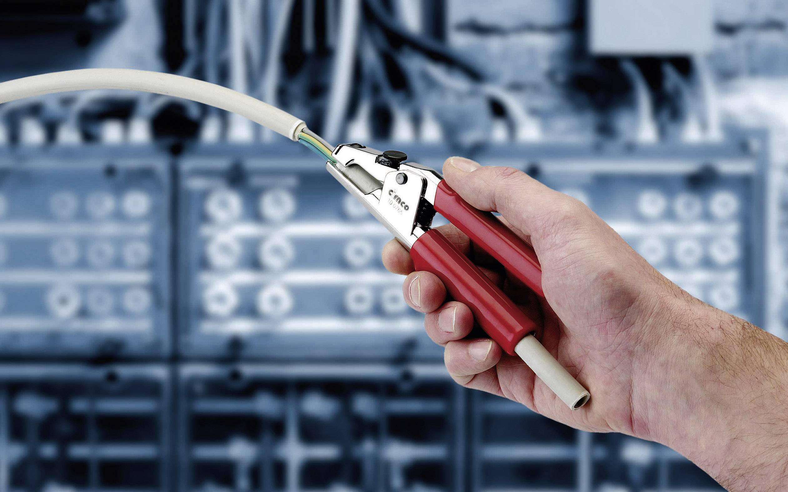 Una mano tiene uno strumento per spellare cavi davanti a uno sfondo sfocato con quadri elettrici.