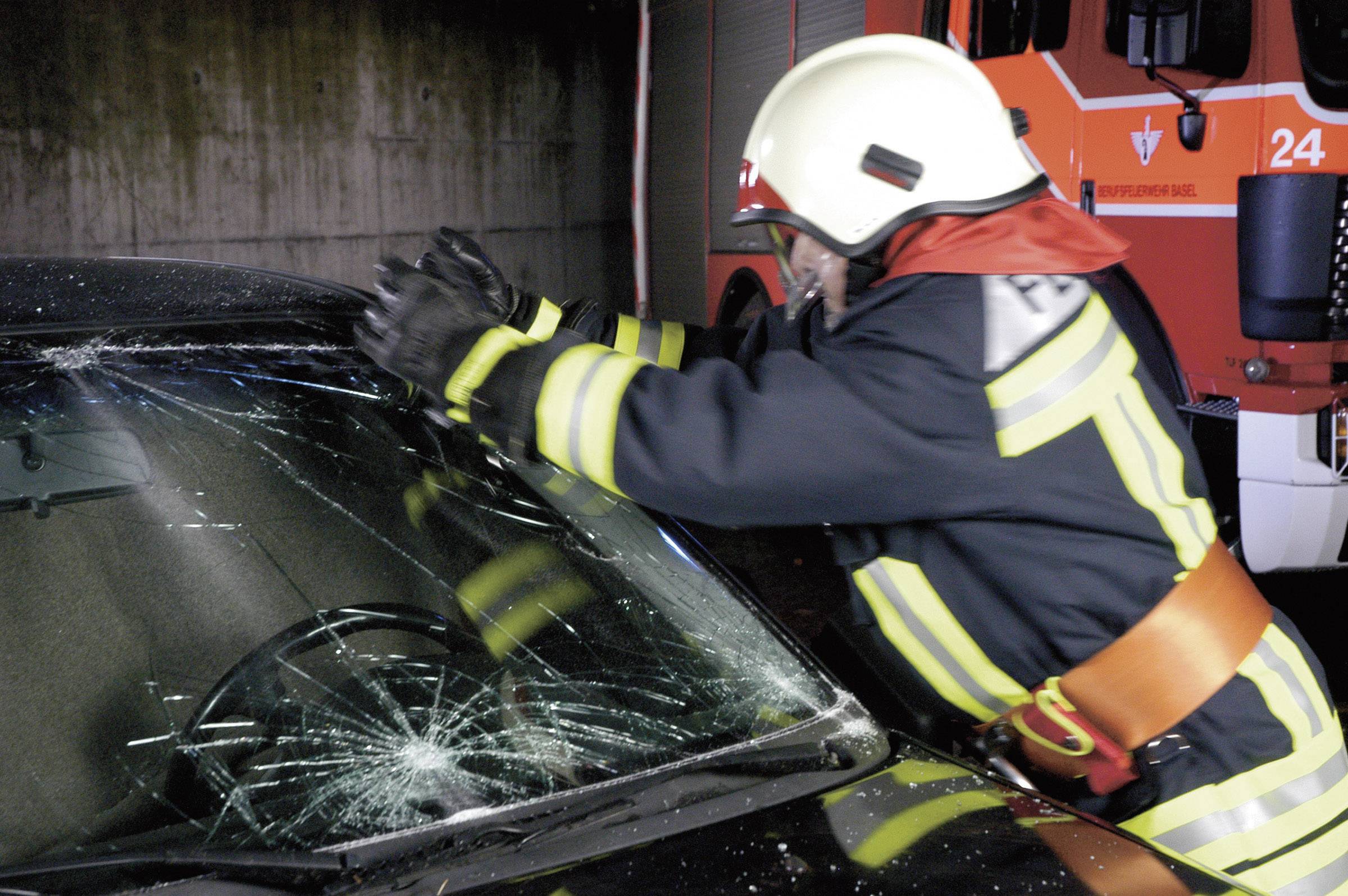 Un vigile del fuoco con indumenti protettivi sfonda il parabrezza di un'auto con un'ascia. Sullo sfondo è presente un'autopompa dei vigili del fuoco.
