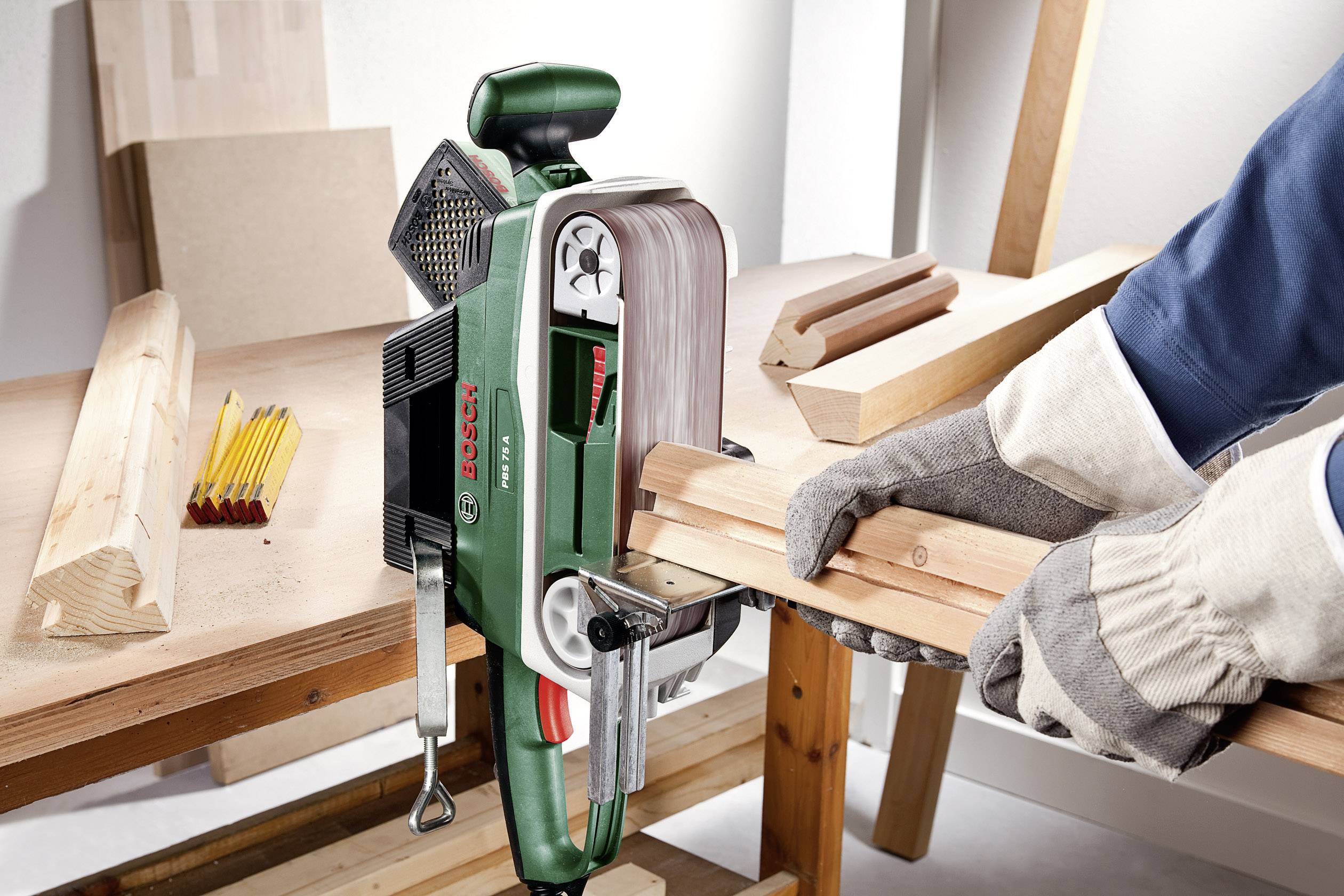 Una persona sta usando una levigatrice per levigare il legno. La levigatrice è verde con dettagli rossi, e sul tavolo ci sono pezzi di legno e matite.