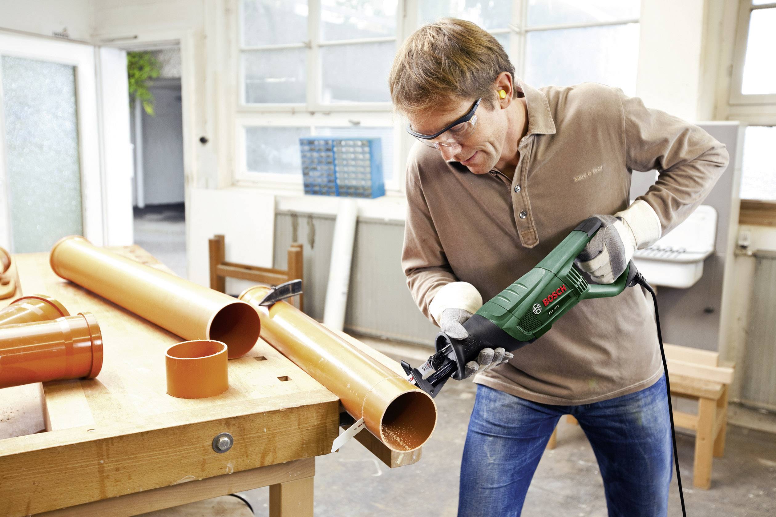 Un uomo con occhiali protettivi e protezioni per l'udito taglia un tubo di plastica arancione con una sega elettrica in un laboratorio.