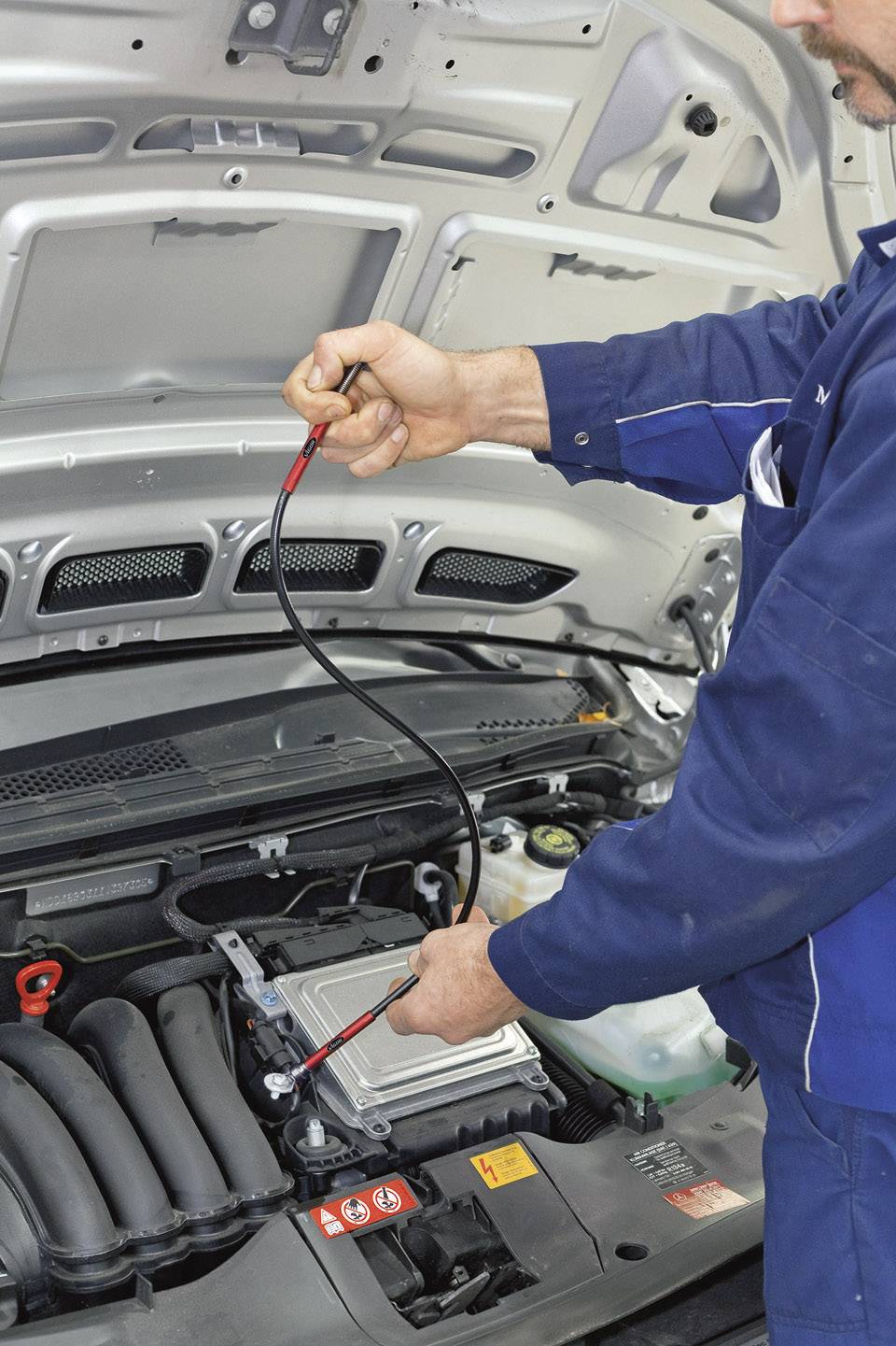 Meccanico in uniforme blu sta controllando il motore di un'auto con uno strumento diagnostico sotto il cofano aperto.