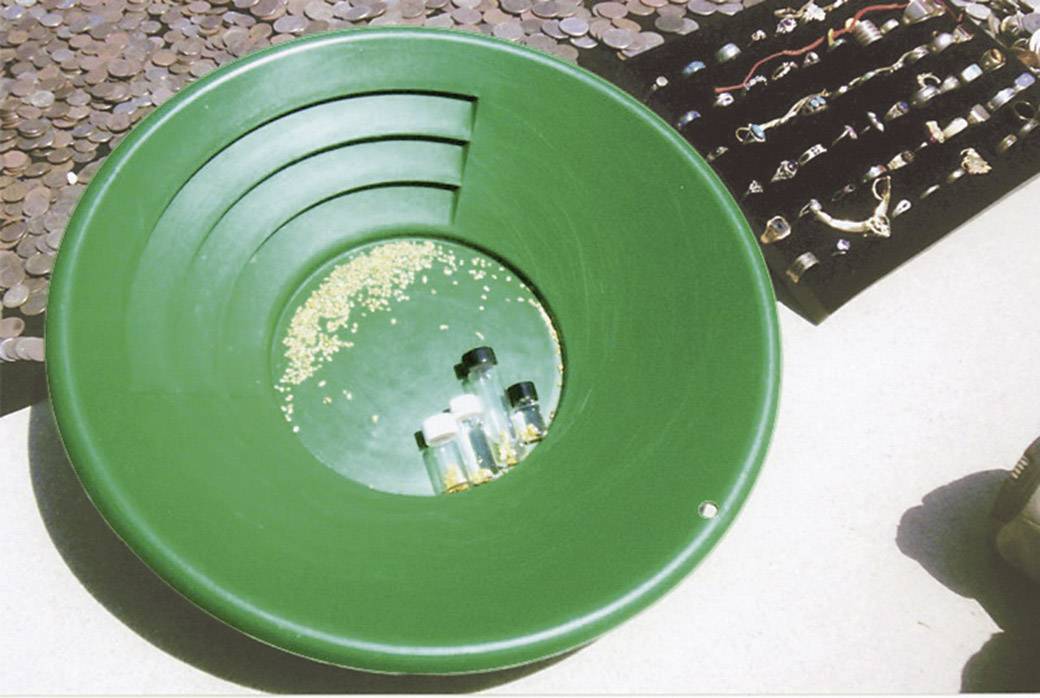 Pentola verde per la ricerca dell'oro con oro raccolto e fiaschetta riempita. Sullo sfondo sono visibili molte monete e gioielli.