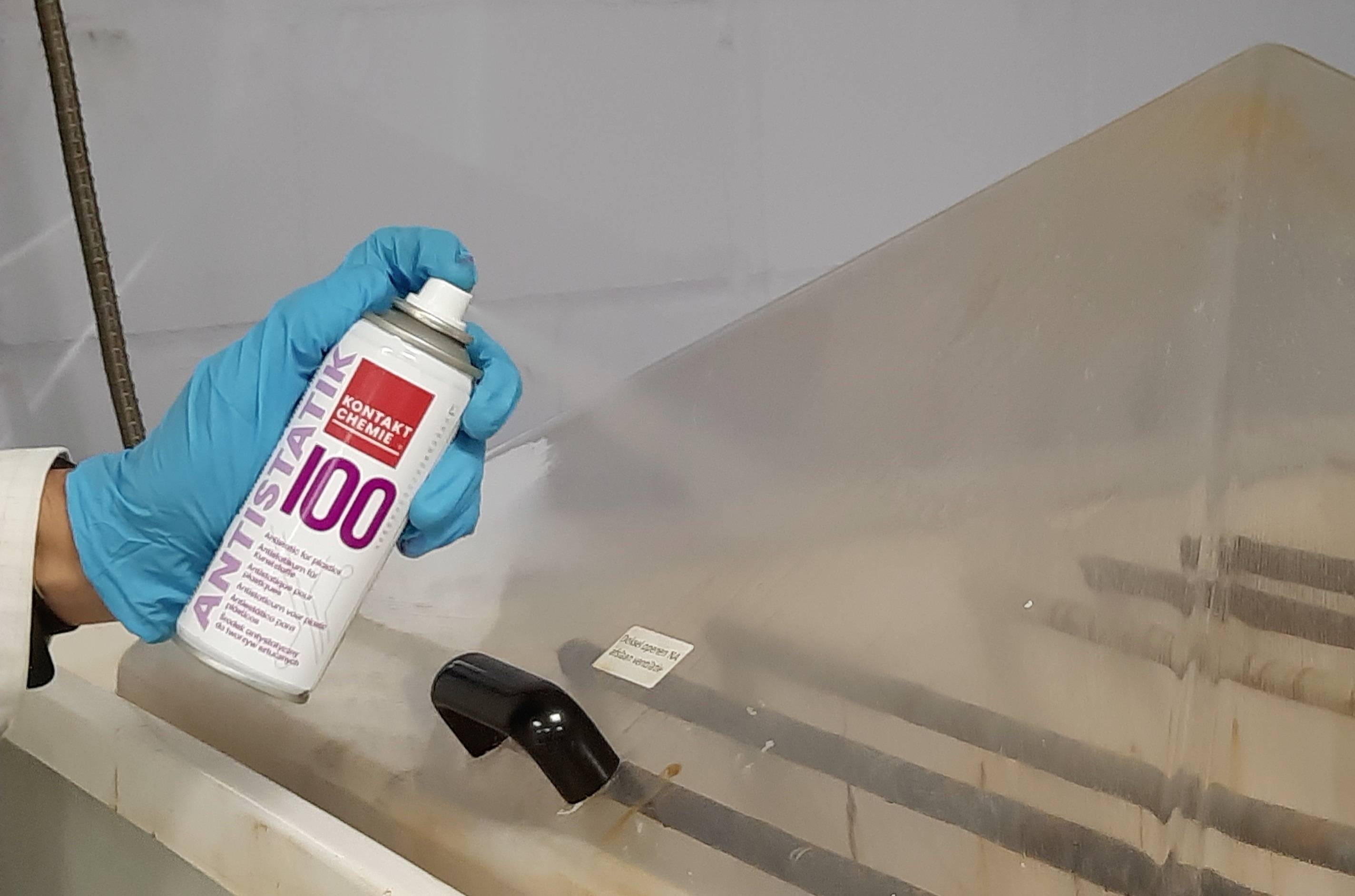 Een gehandschoende hand houdt een anti-statische spuitbus vast in de buurt van een grote afzuigkap in een laboratoriumomgeving.