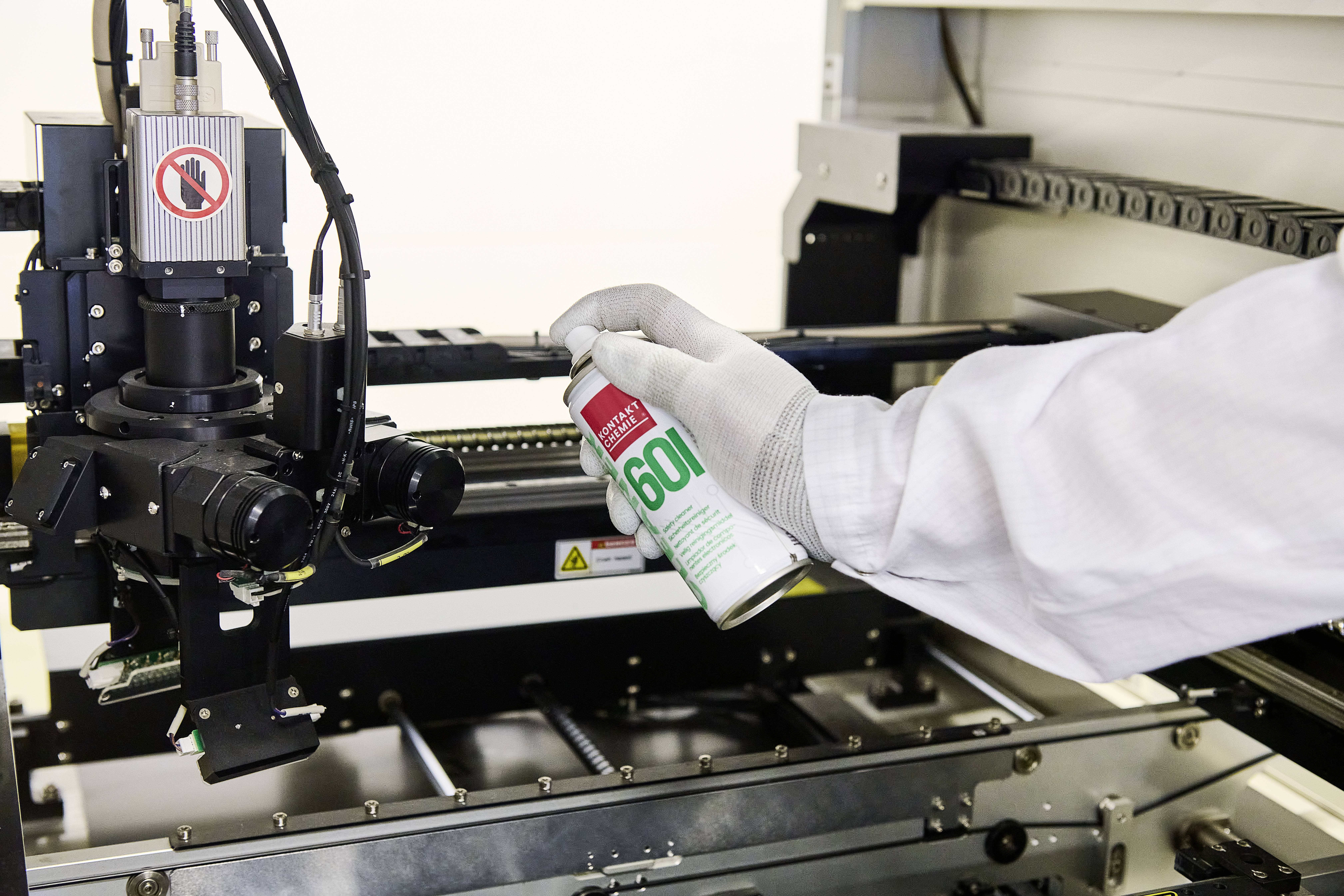 Een persoon met handschoen besproeit een machine met een spray voor reiniging of onderhoud. Een verbodsbord is zichtbaar.
