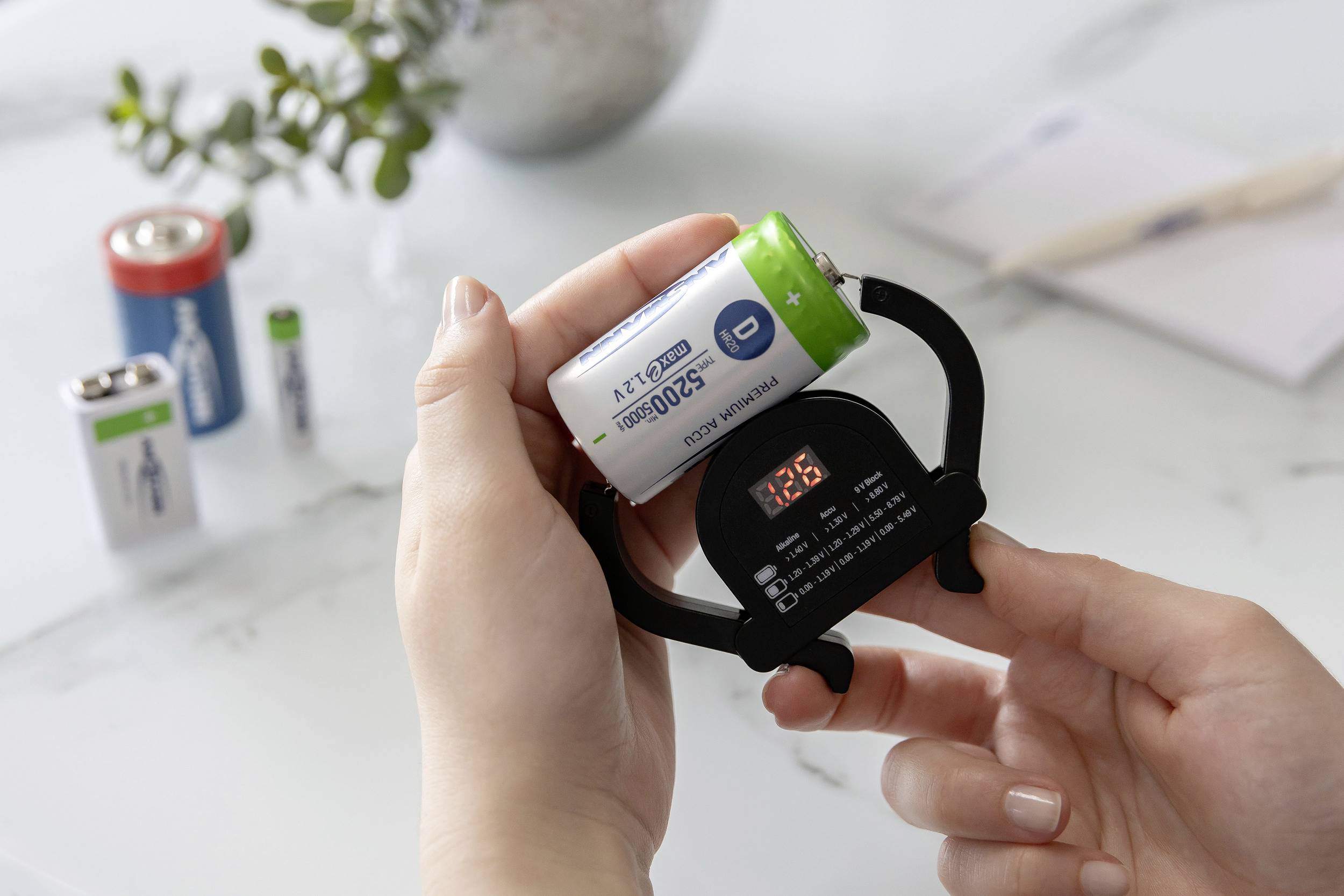 'Persoon meet de laadstatus van een batterij met een batterij-tester in de hand; meer batterijen op de achtergrond op tafel.'