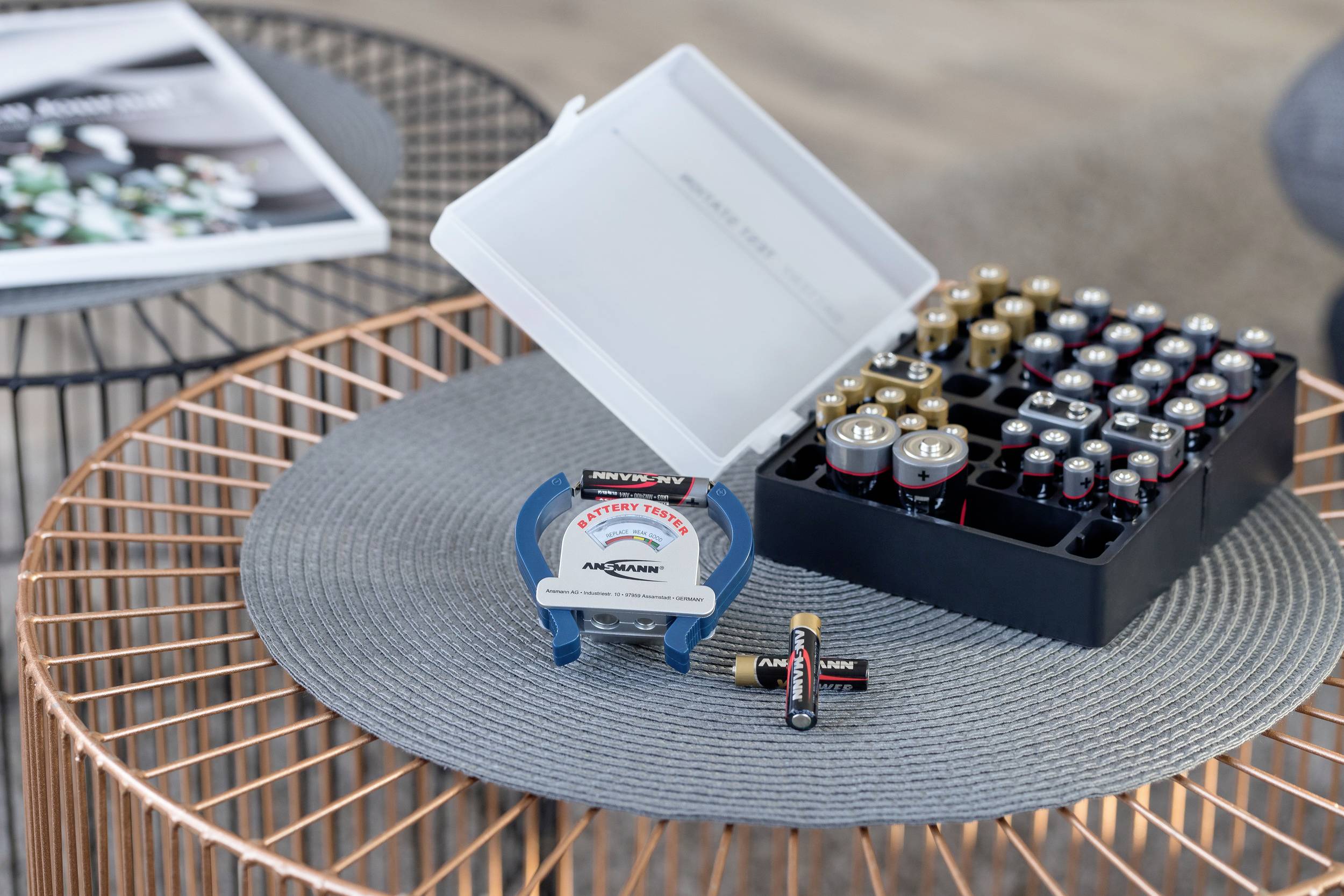 'Een tafel met een rond kleed waarop een batterijentester staat, batterijen in een doos en enkele losse batterijen in de buurt.'