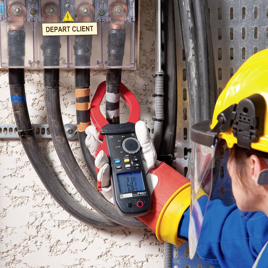 Een persoon in werkkleding meet de elektrische stroom aan een schakelkast met een multimeter. Veiligheidsuitrusting wordt gedragen.