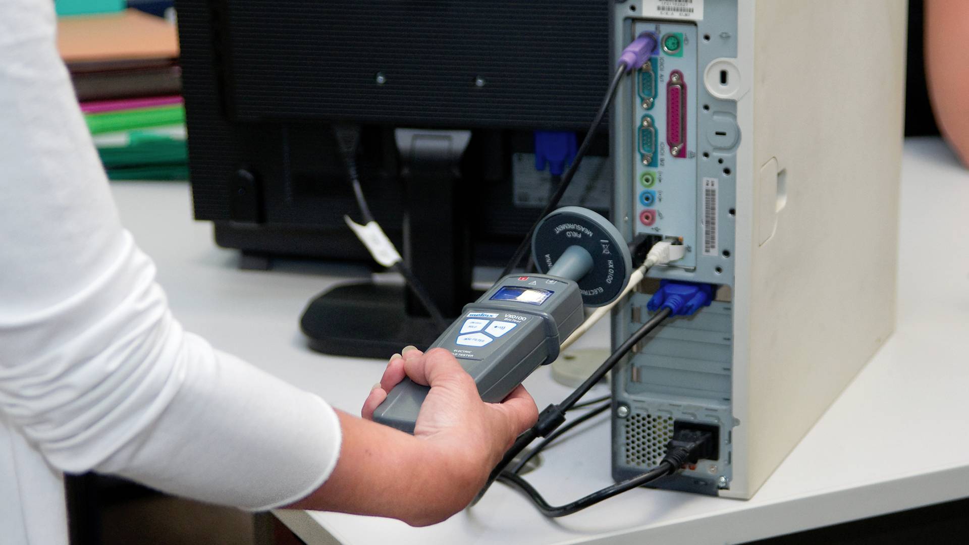 Een persoon houdt een elektrisch meetapparaat tegen de achterkant van een geopend computerbehuizing om metingen aan de aansluitingen uit te voeren.