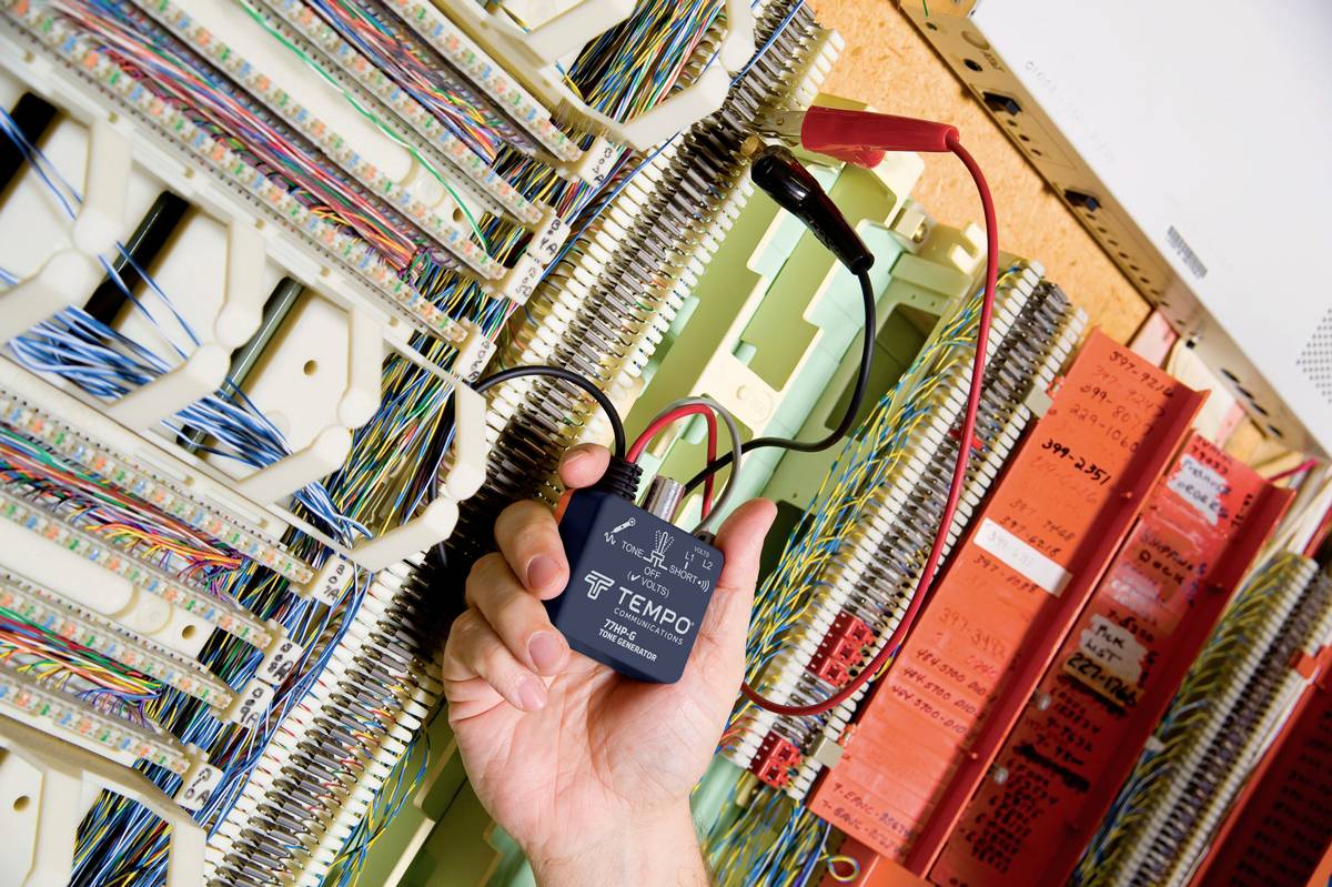 'Een persoon houdt een zwart apparaat met kabels in de hand, omringd door talrijke telefoonstrips in een schakelkast.'