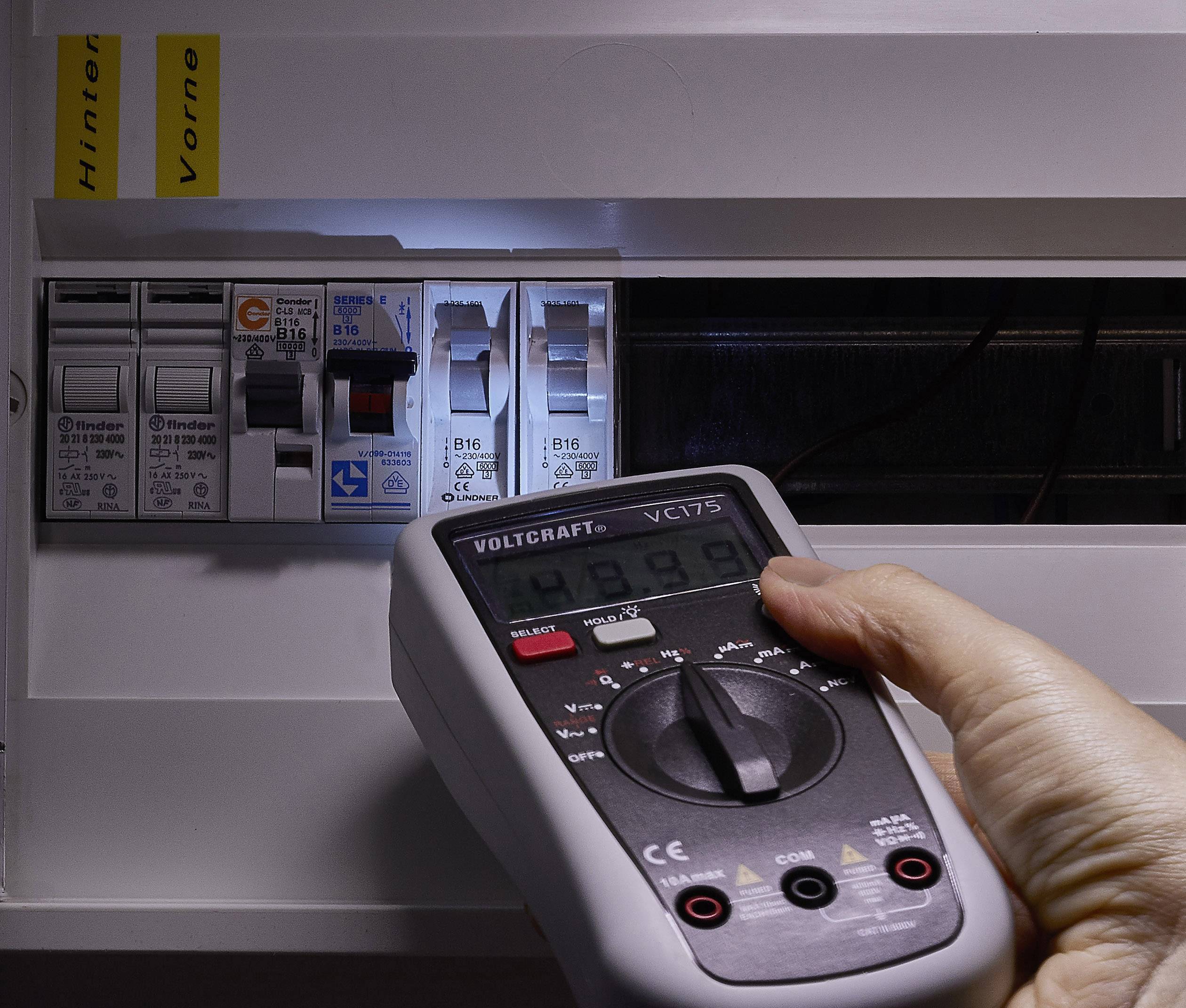 Hand houdt een multimeter voor een geopende zekeringkast, controleert de spanningswaarden. De focus ligt op precisie en veiligheid.