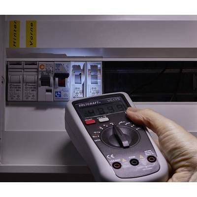 Hand houdt een multimeter voor een geopende zekeringkast, controleert de spanningswaarden. De focus ligt op precisie en veiligheid.
