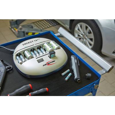 Batterijlader op werkbank met opgeladen batterijen, gereedschappen ernaast. Auto op de achtergrond. Werkplaatsomgeving.