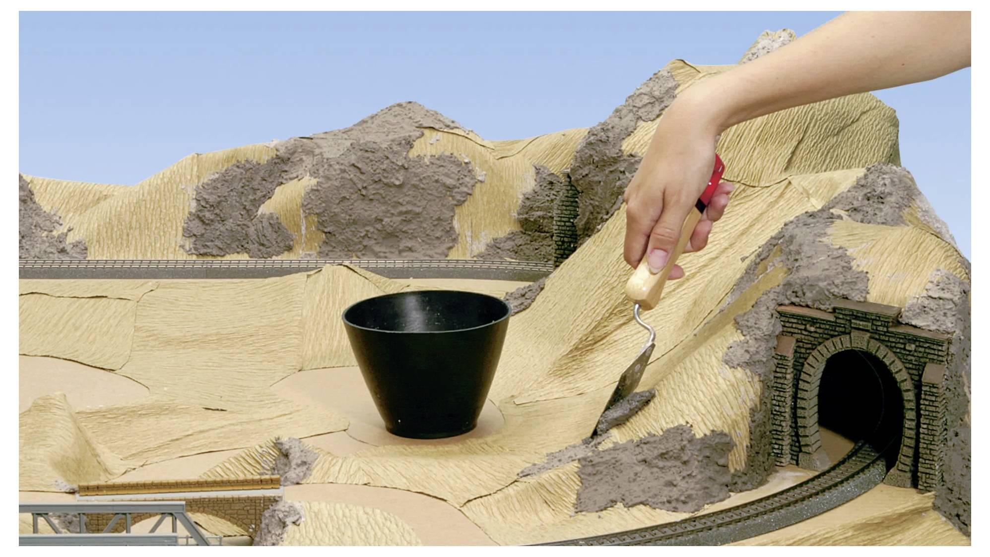Een modellandschap dat een hand toont die een scultuurgereedschap gebruikt om terreindetails in een dioramaopstelling met een tunnel en sporen uit te snijden.