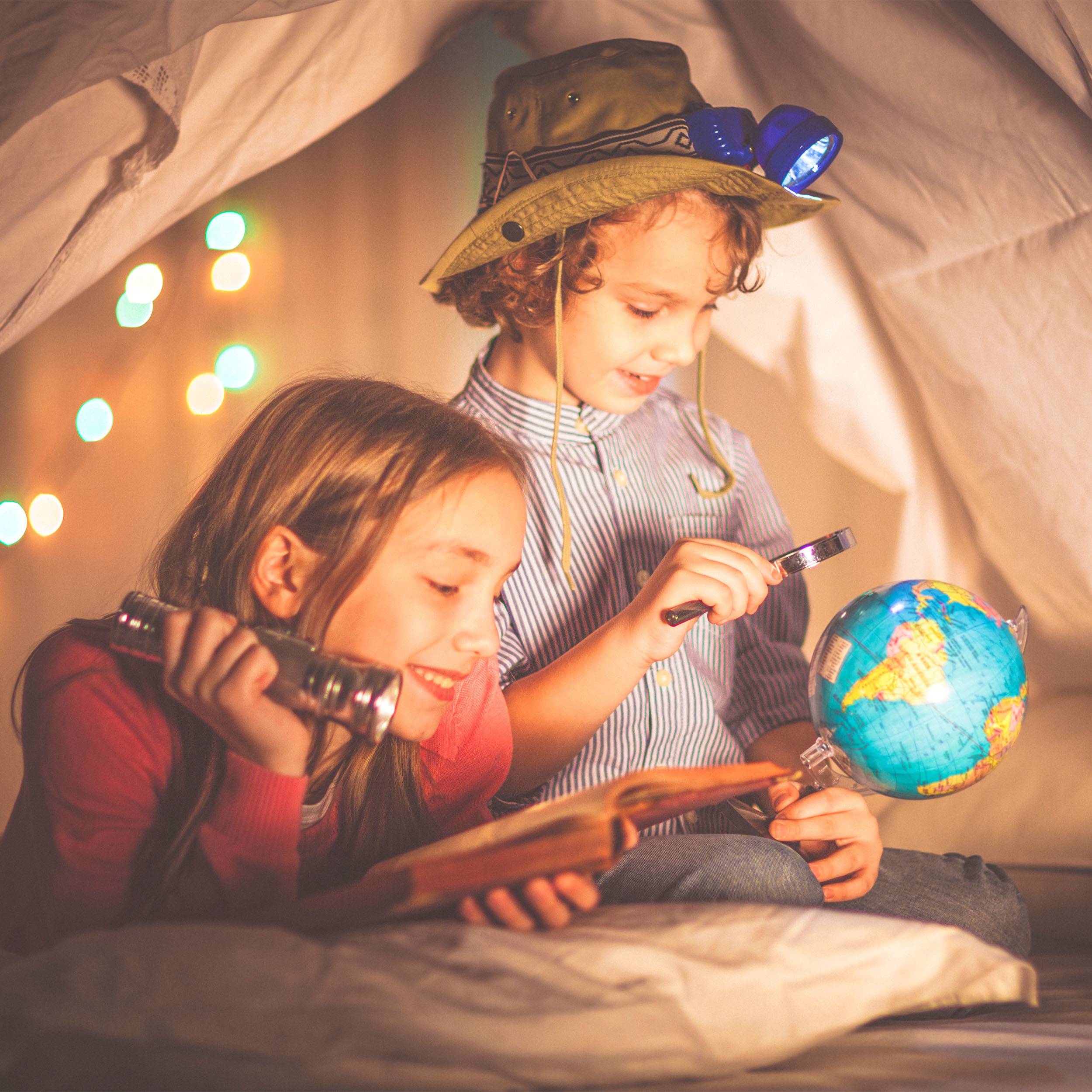 Twee kinderen liggen in een tent, nieuwsgierig een boek aan het bekijken. Eén houdt een verrekijker vast, de ander wijst naar een globe.