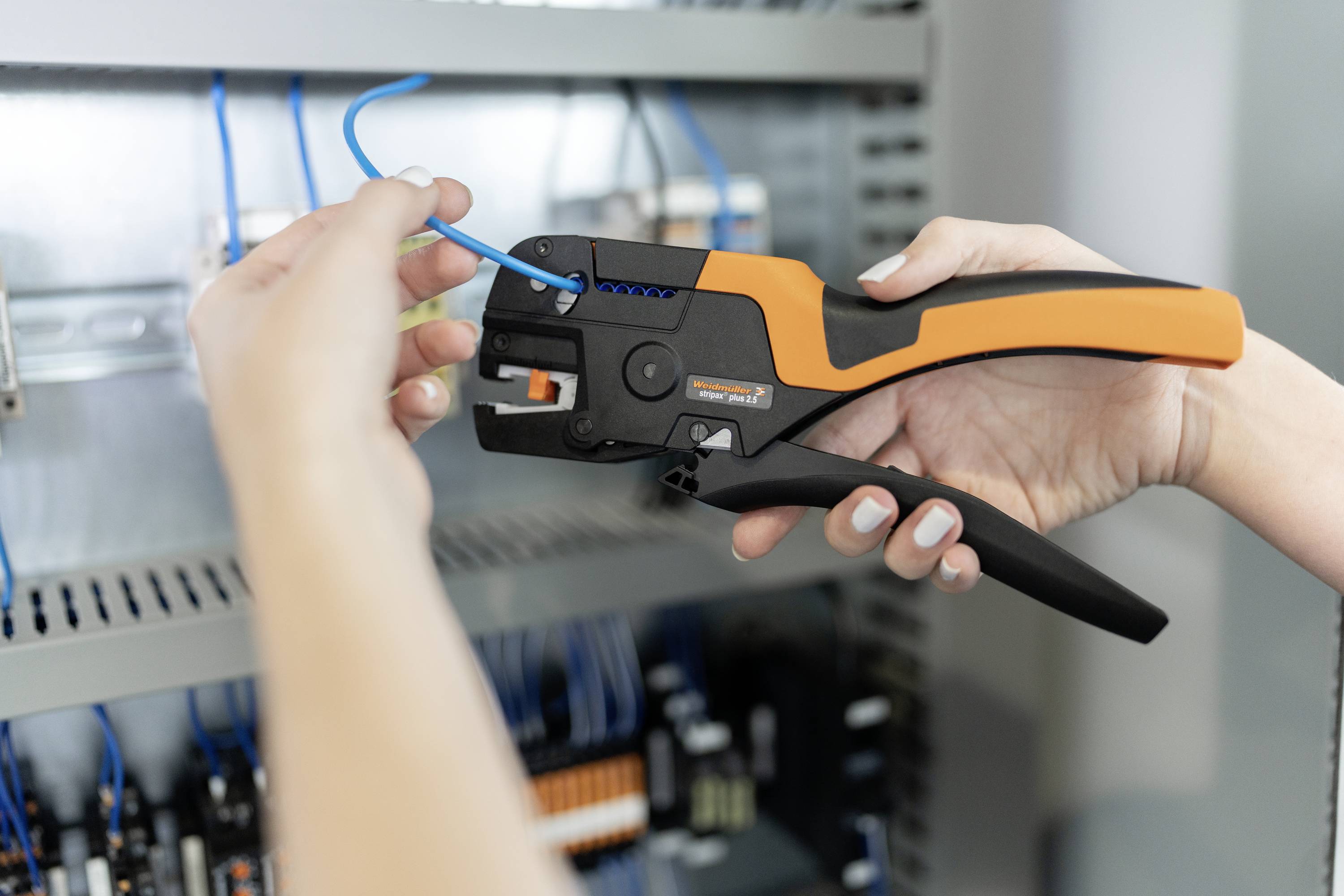 Een persoon gebruikt een oranje-zwart gereedschap om een blauw kabel in een elektriciteitskast te bewerken.
