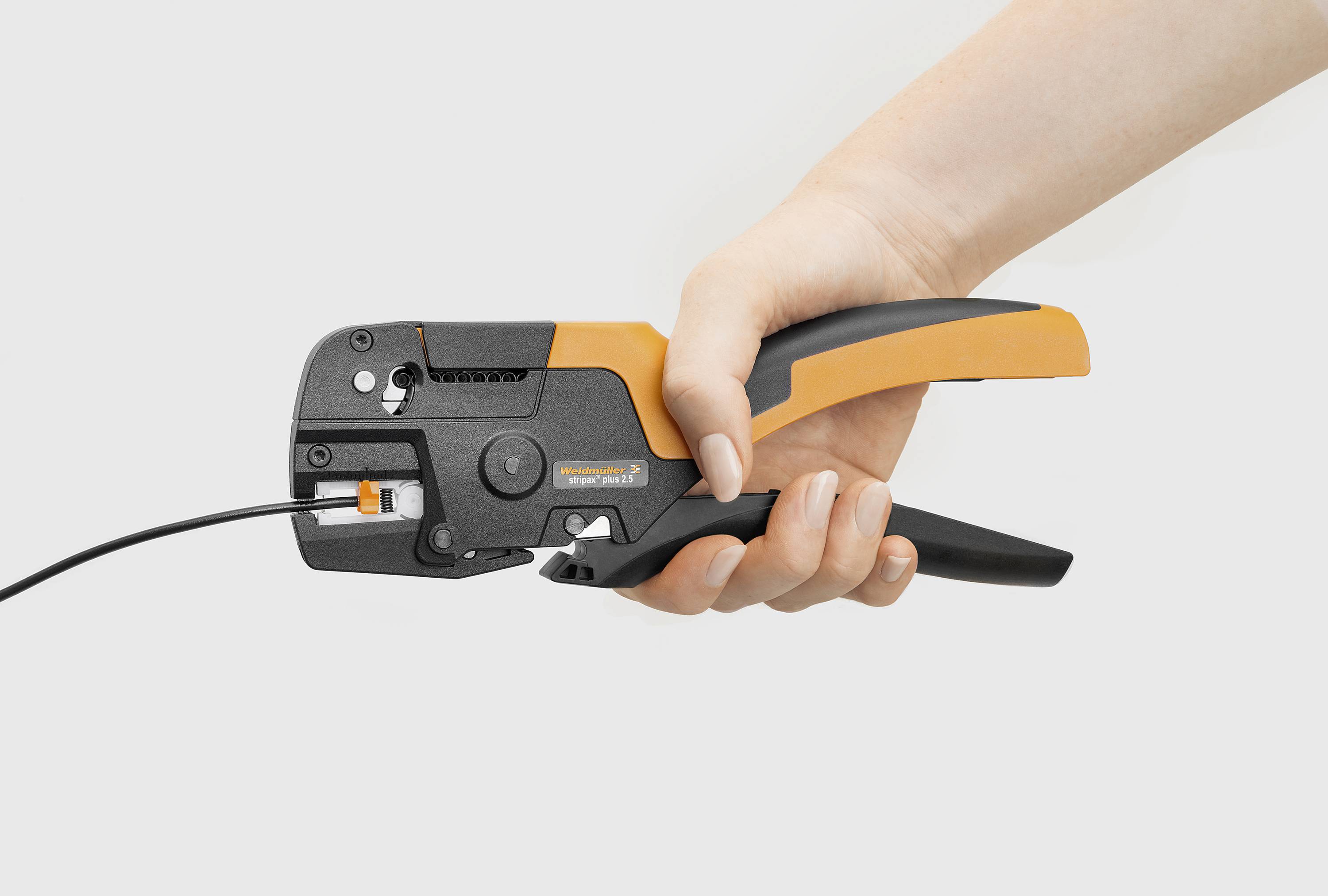 Een hand houdt een oranje-zwarte krimptang vast die een kabel bewerkt, voor een neutrale achtergrond.