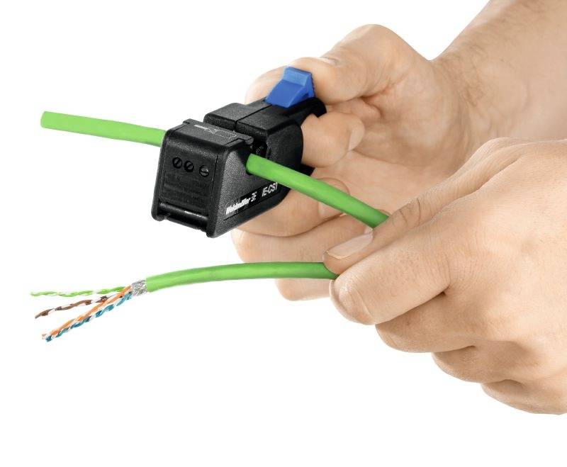 Handen houden een kabelstripper vast die een groen kabel ontmantelt. De binnenste draden van de kabel zijn zichtbaar.