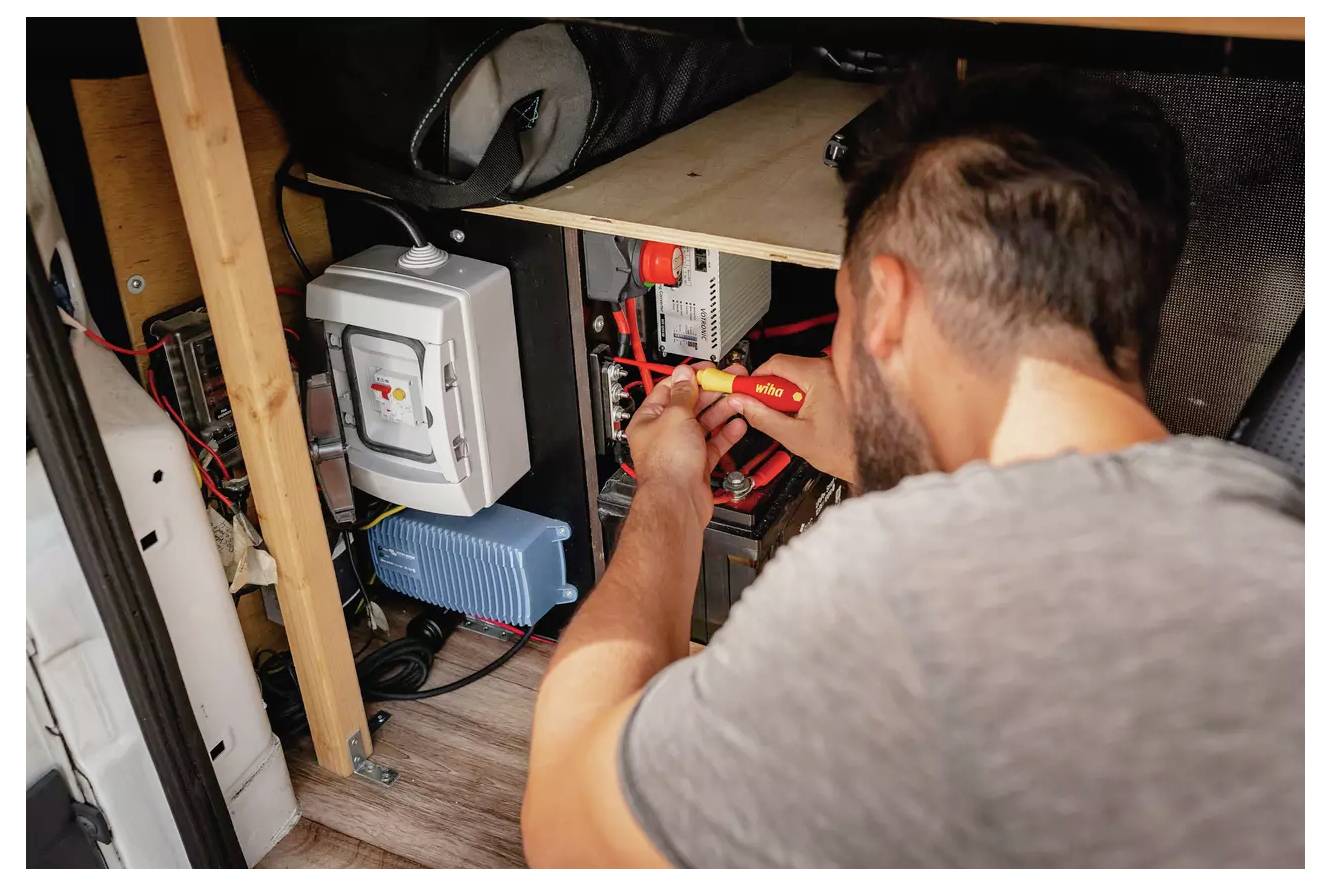 Een persoon maakt met een schroevendraaier draadverbindingen vast in een kastje met elektrische componenten, inclusief een blauw apparaat en zekeringautomaten.