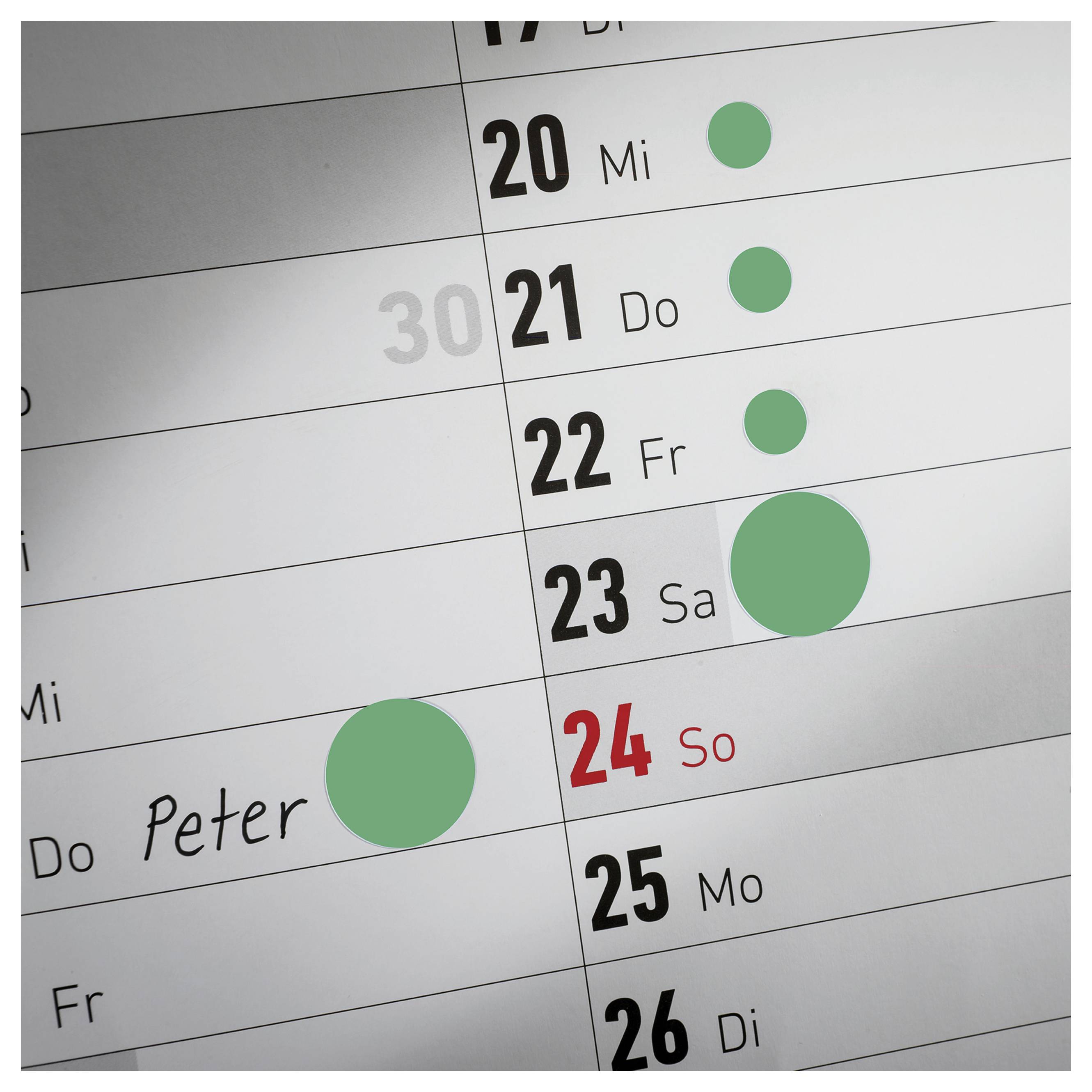 Close-up van een wandkalender met datum 20 tot 26; groene punten markeren afspraken, op 24. zondag is een afspraak rood gemarkeerd.