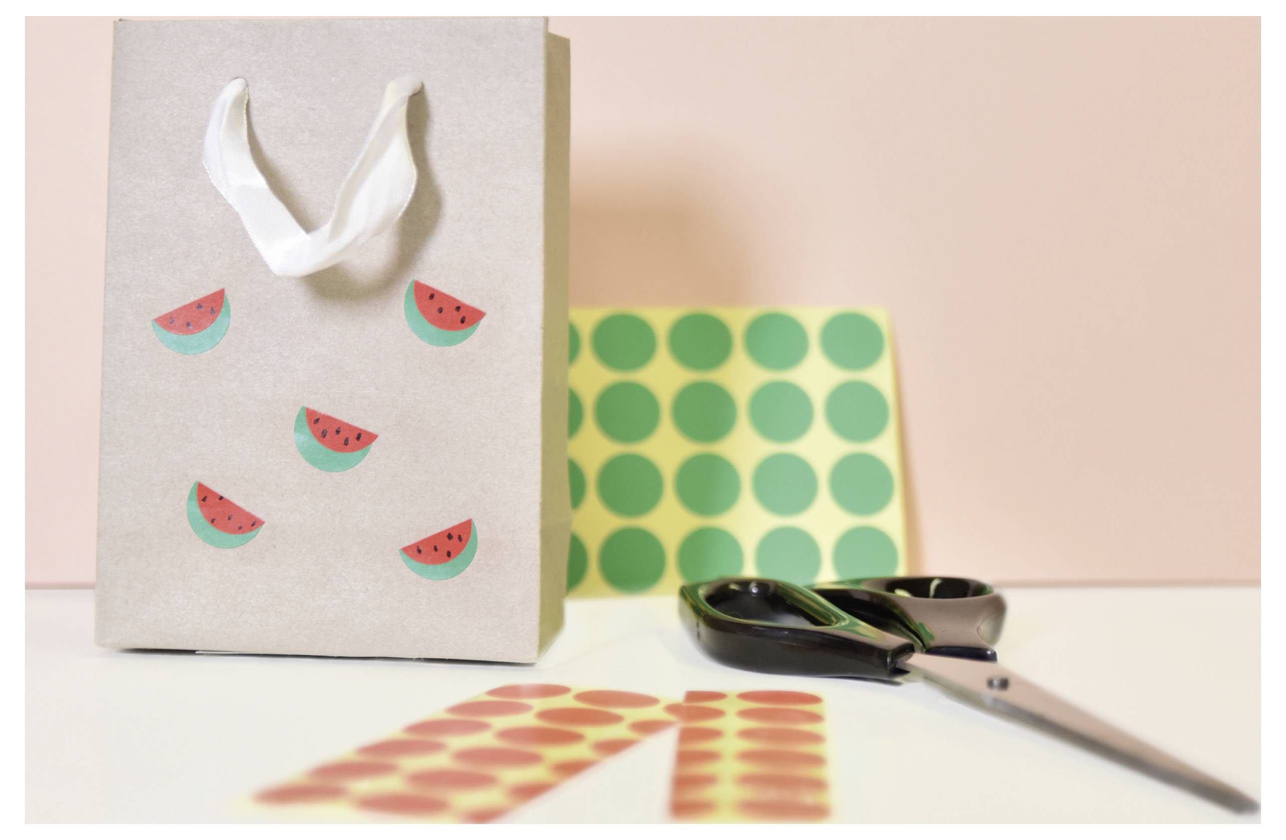 Cadeauzakje met watermeloen-stickers naast schaar en gemusterd papier voor knutselen en inpakken op een lichte tafel.
