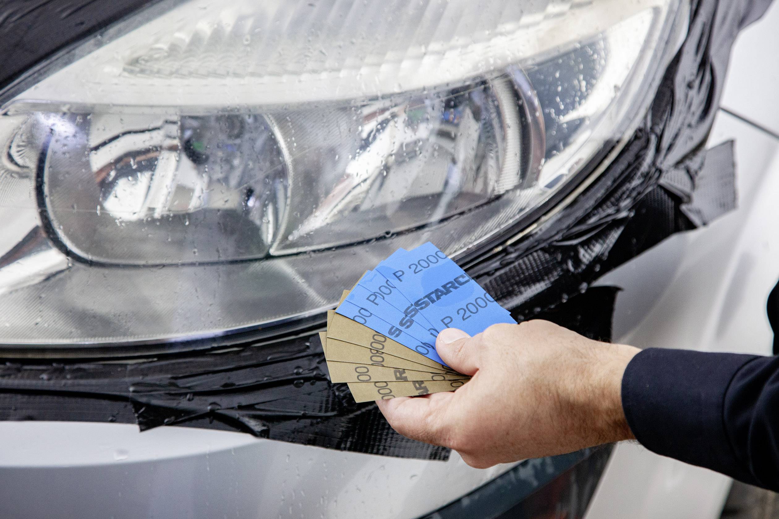 Een hand houdt verschillende schuurpapieren voor een natte autokoplamp die met zwart plakband is omwikkeld.