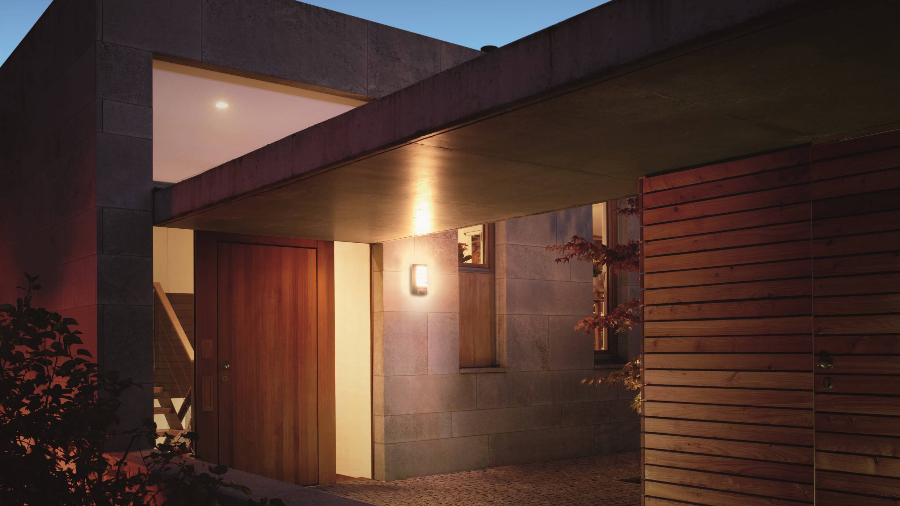 Een modern huis bij schemering met ingeschakelde buitenverlichting. De voordeur is van hout, de gevel is van steen en hout.