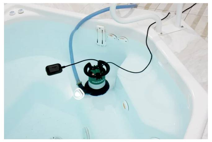 Een pomp ondergedompeld in een met water gevuld bad, verbonden met een blauwe slang en zwart stroomsnoer, wat duidt op afvoer of reiniging van het bad.