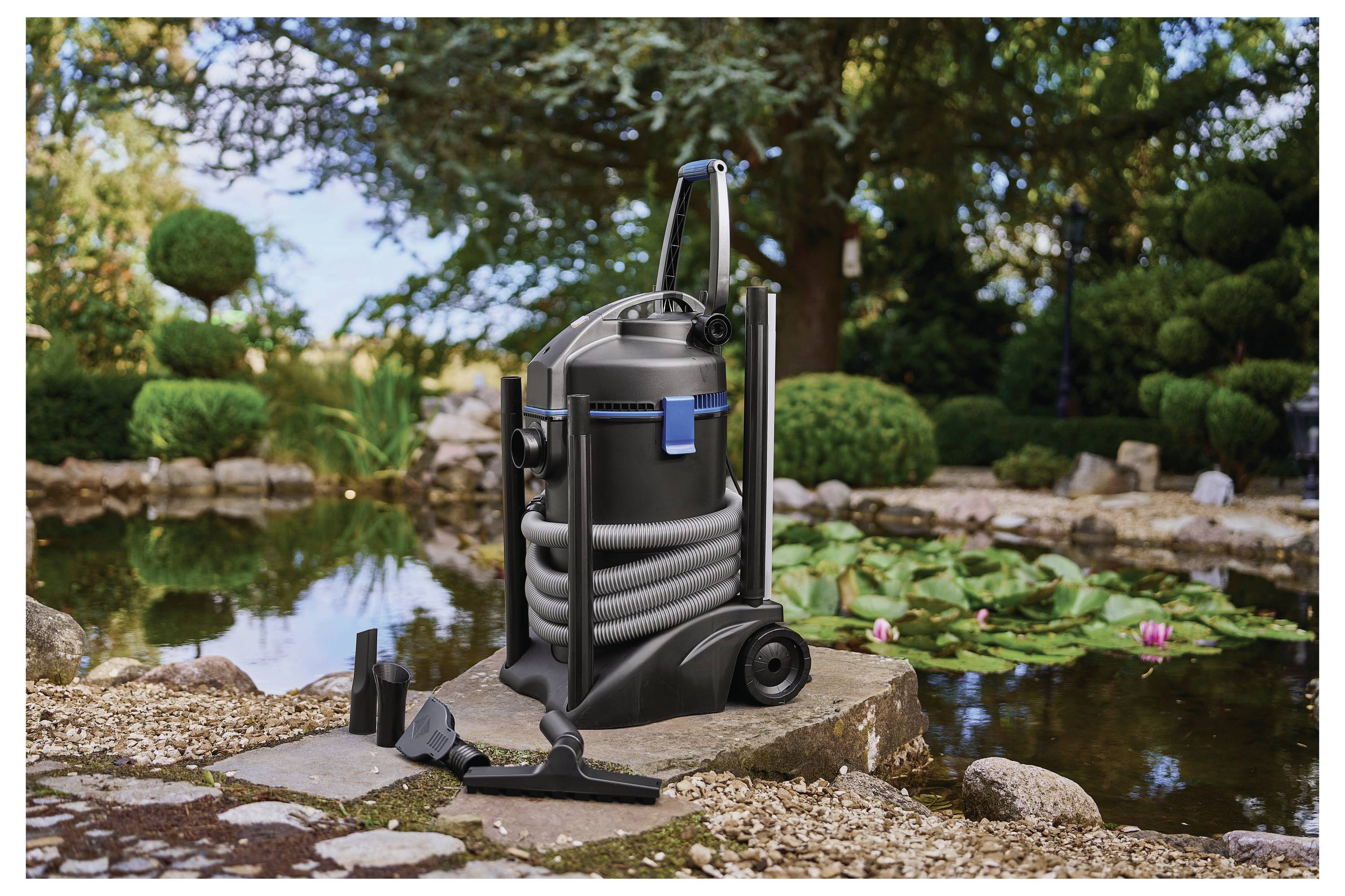 Een vijverstofzuiger op een stenen pad naast een tuinvijver met waterlelies, rotsen en weelderig groen op de achtergrond.