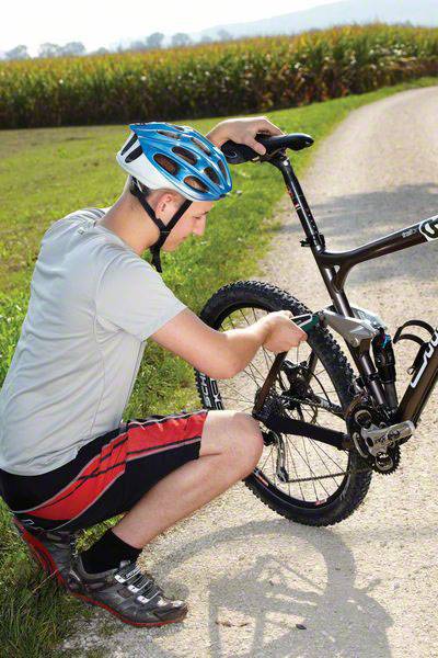 Een persoon draagt een helm en knielt op een veldweg terwijl hij het achterwiel van een fiets repareert. Op de achtergrond is een veld te zien.