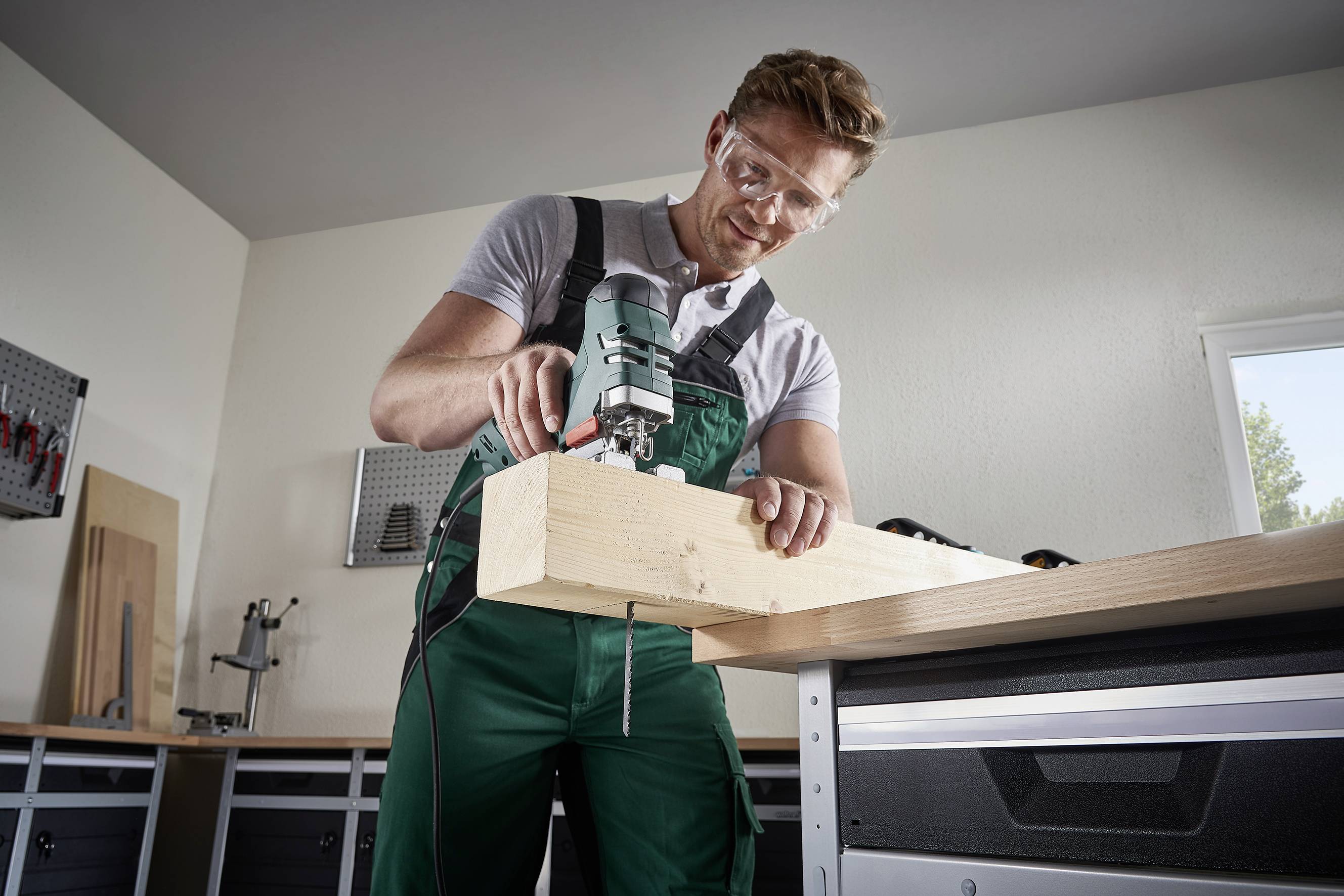 Een man bewerkt een houten stuk in een werkplaats met een elektrische zaag. Hij draagt een veiligheidsbril en werkkleding.