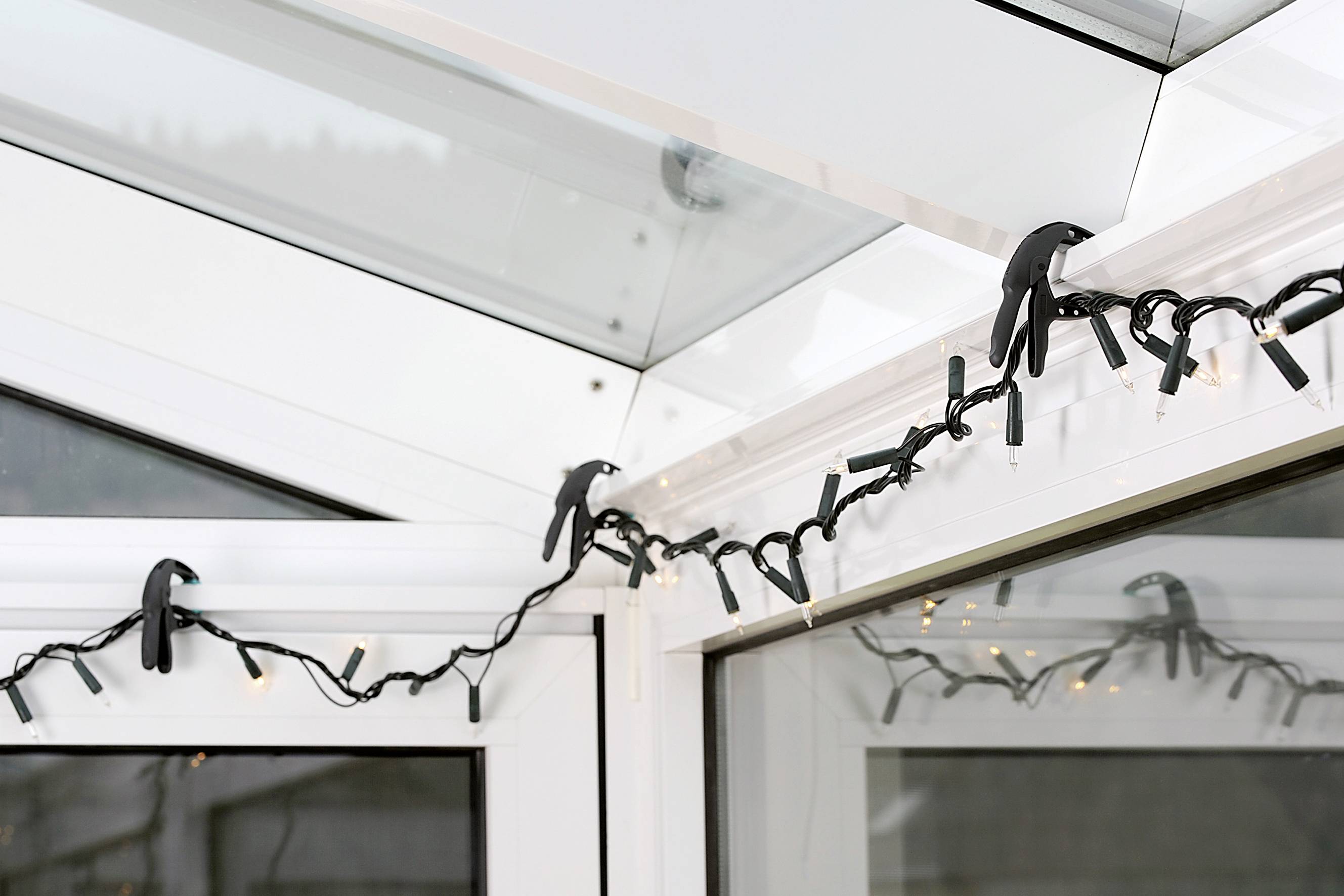 Een lichtslinger is langs de hoek van een witte wintertuin aangebracht, met clips aan het frame bevestigd. Enkele lampen zijn ingeschakeld.