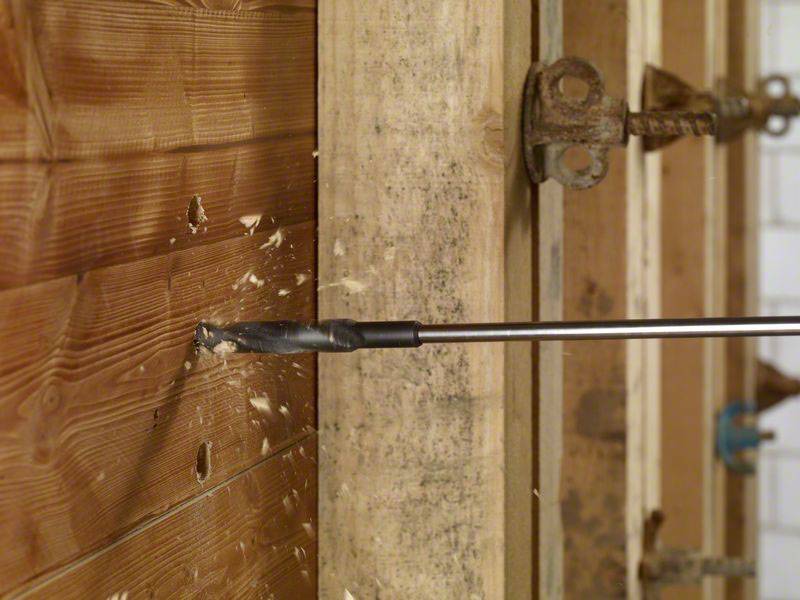 Een boor dringt door een houten wand in een werkplaats- of bouwomgeving, houtspaanders vliegen rond. Een spaninrichting en gereedschap zijn op de achtergrond zichtbaar.