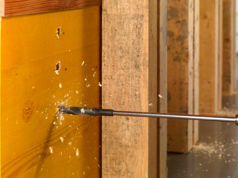 Een boormachine boort door een houten muur, houtspaanders vliegen rond. Duidelijk zichtbare geboorde gaten tonen de voortgang van het werk.
