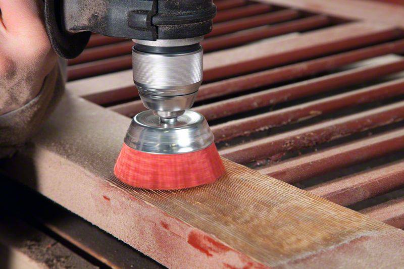 Een persoon schuurt hout met een boormachine en een borstelopzetstuk. Het hout heeft een rechthoekige vorm met meerdere sleuven.