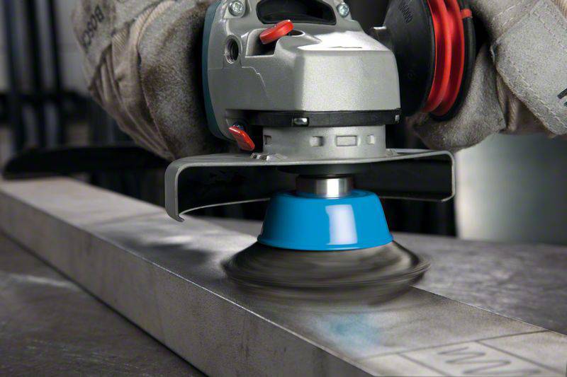 Een polijstmachine met een roterende schuurkop bewerkt het oppervlak van een metalen strip, gedragen door handen in werkhandschoenen.