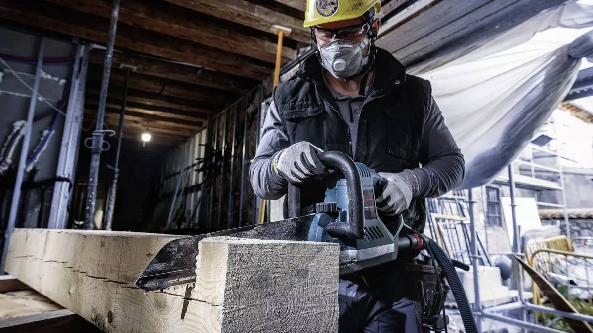 Een bouwvakker met veiligheidsmasker en helm zaagt een houten balk in een werkplaats. Hij gebruikt een elektrische zaag.