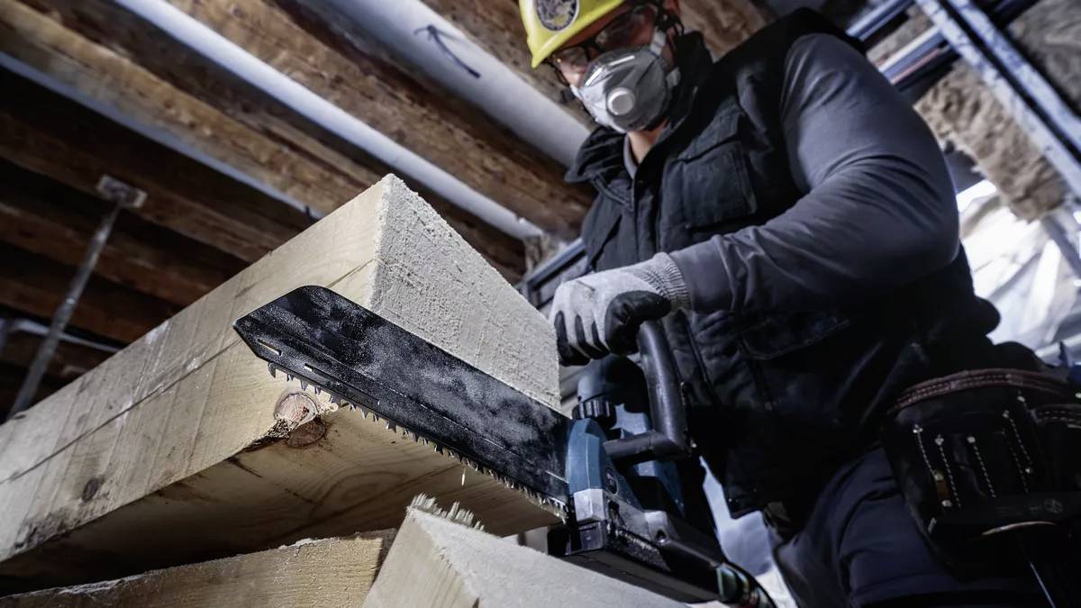 Een bouwvakker met een veiligheidshelm en masker zaagt hout met een kettingzaag in een casco. Boven zijn onbedekte houten balken zichtbaar.