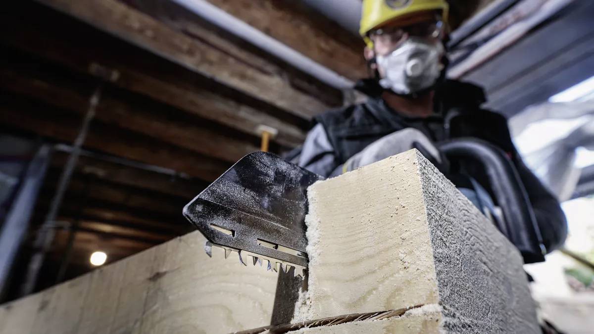 Een bouwvakker met veiligheidsbril en ademhalingsmasker zaagt een houten plank in een werkplaats.