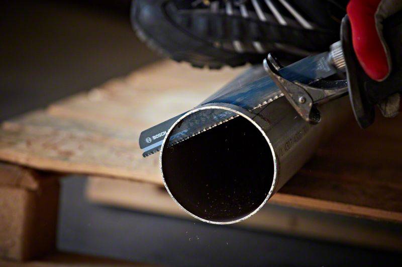 Een persoon zaagt een metalen buis door met een handzaag. De buis ligt op een houten pallet. Vonken spatten rond tijdens het zagen.