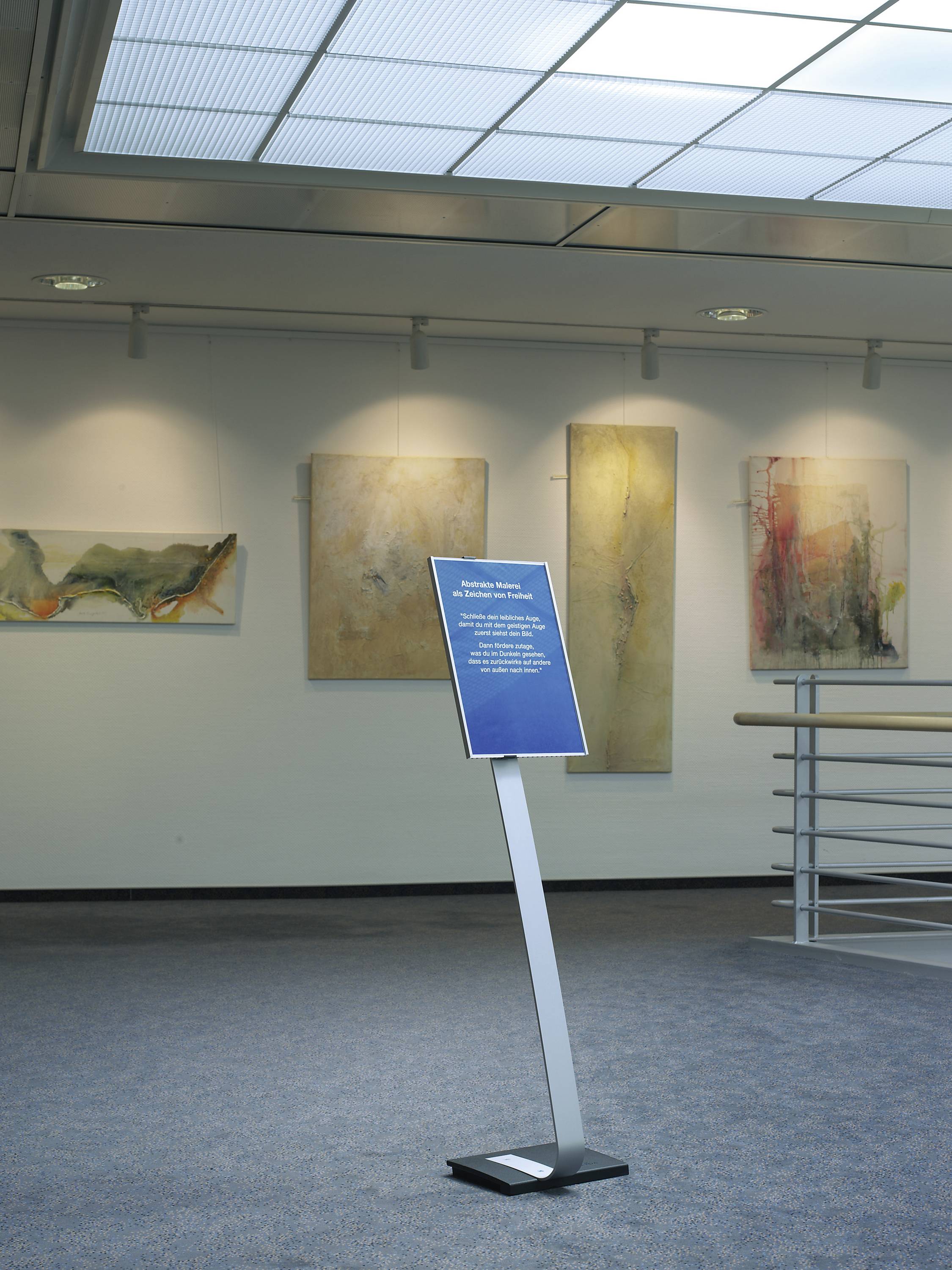 Een bordje staat in een galerij met abstracte schilderijen aan de muur. De omgeving oogt modern en minimalistisch.