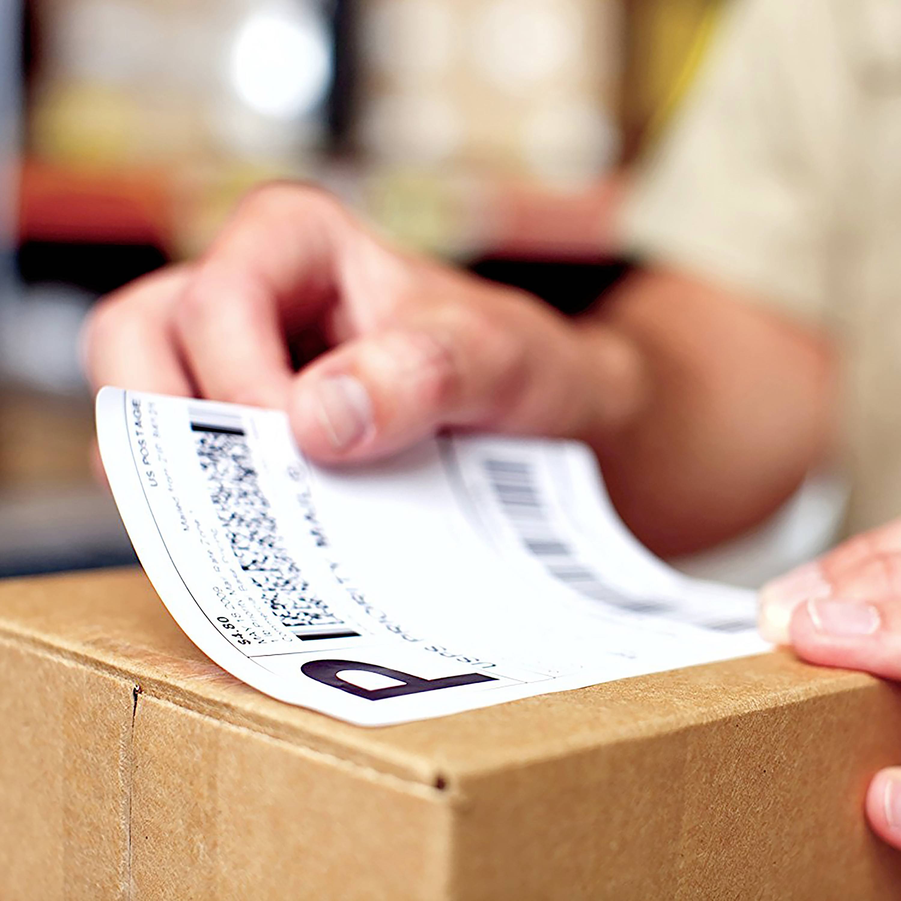Een hand houdt een verzendingsetiket vast op een bruine verpakking. De achtergrond is wazig.