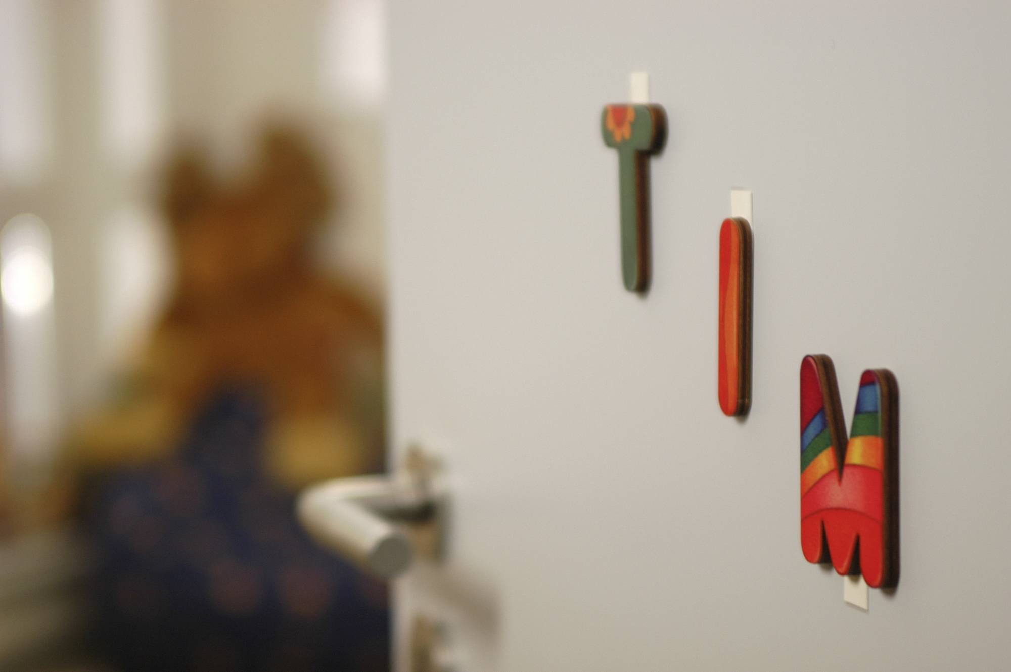 Deur met gekleurde houten letters 'T I M'; onscherpe achtergrond toont een glimp van een kinderkamer met knuffel.