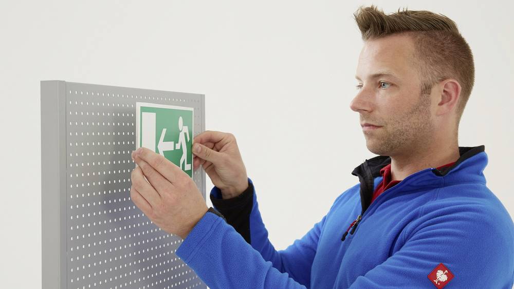 Een man in een blauwe jas bevestigt een groen nooduitgangsbord aan een geperforeerde plaat.