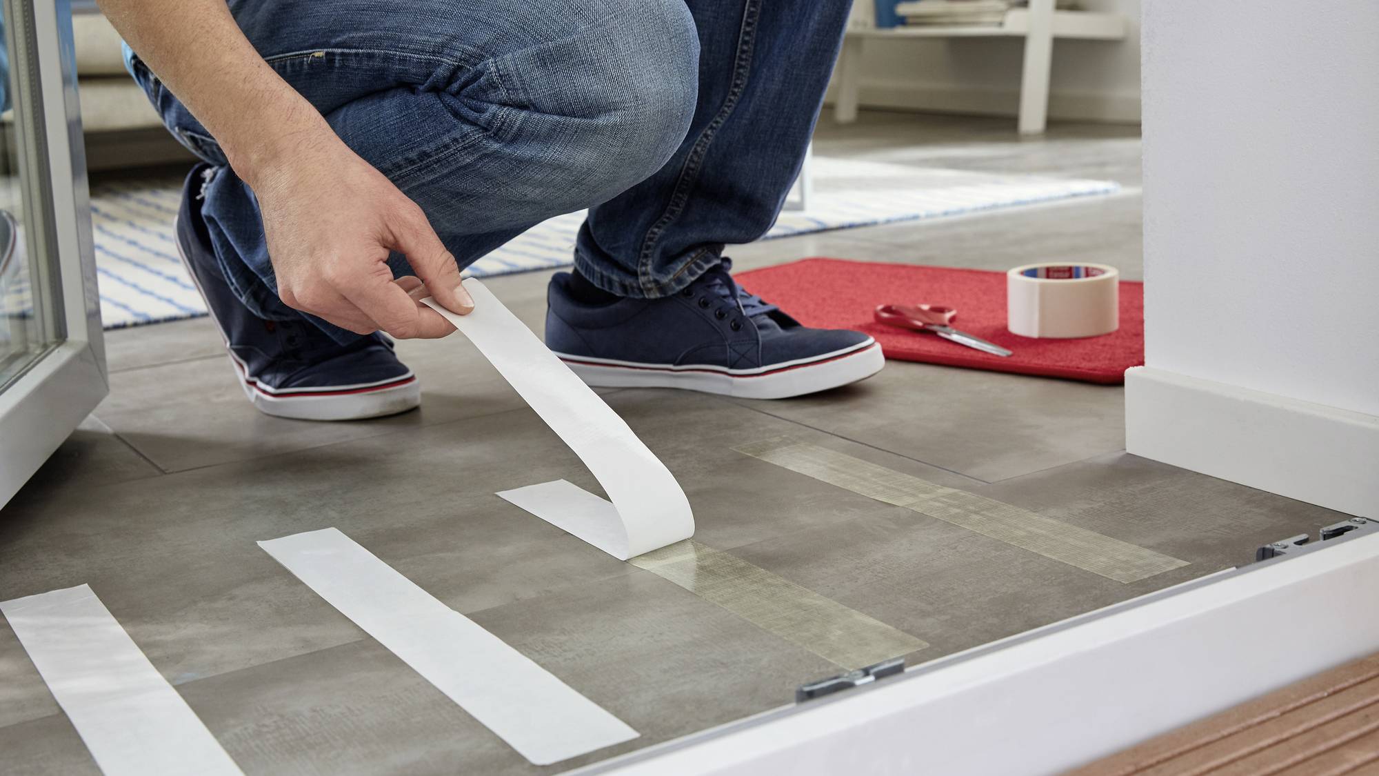 Een persoon plakt witte antislipstrips op een tegelvloer om uitglijden te voorkomen. Op de achtergrond ligt rood plakband.