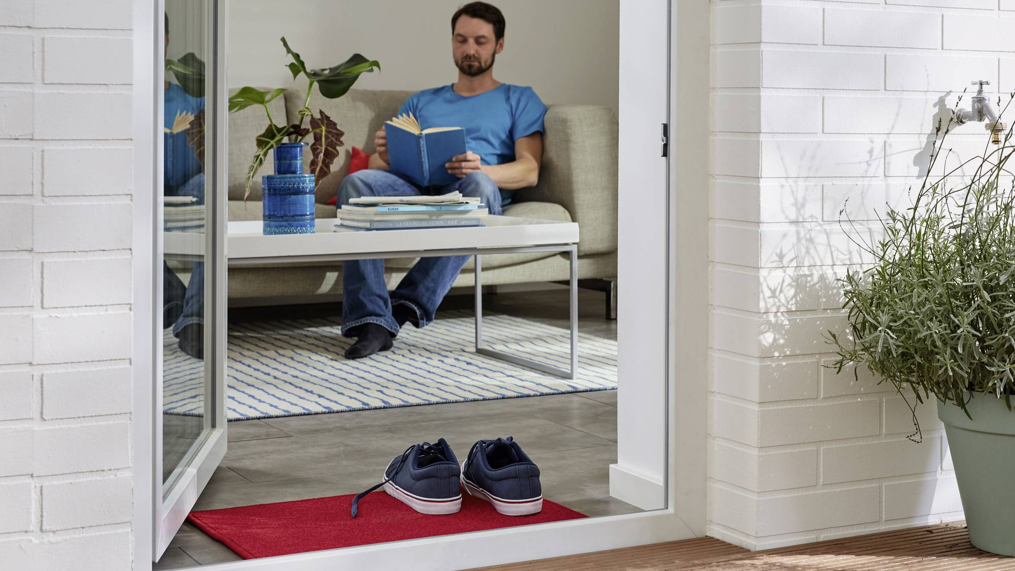 Een man zit te lezen op een bank in de woonkamer. Op de voorgrond liggen een paar schoenen op een rood tapijt voor een open deur.