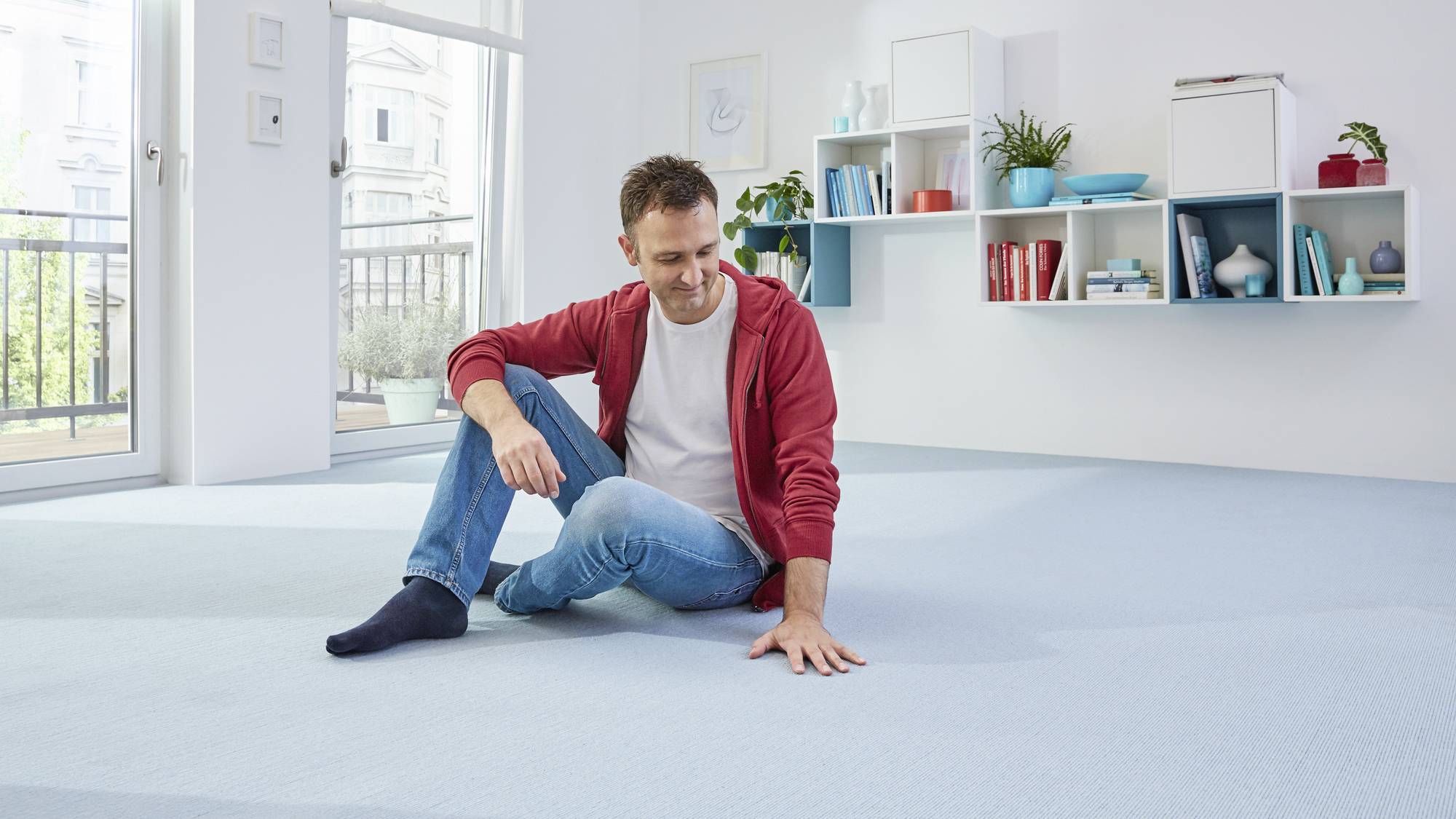 Een man zit glimlachend op een lichtblauwe vloerbedekking in een modern ingericht woonkamer met planken op de achtergrond.