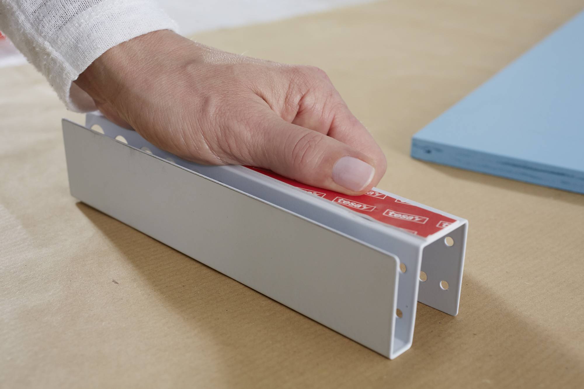 Een hand houdt een U-vormig metalen stuk vast met rode plakband, dat op een tafel ligt. Ernaast ligt een blauw rechthoek.