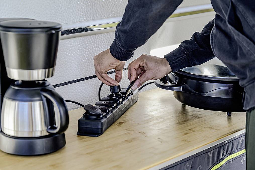 Een persoon sluit een elektrisch apparaat aan op een stekkerdoos in een keukenblok. Een koffiezetapparaat en kookplaat zijn zichtbaar.