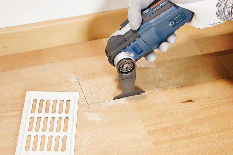 Een ambachtsman gebruikt een elektrisch zaaggereedschap om een rechthoekige vorm in een houten vloer te zagen. Een ventilatieafdekking ligt ernaast.